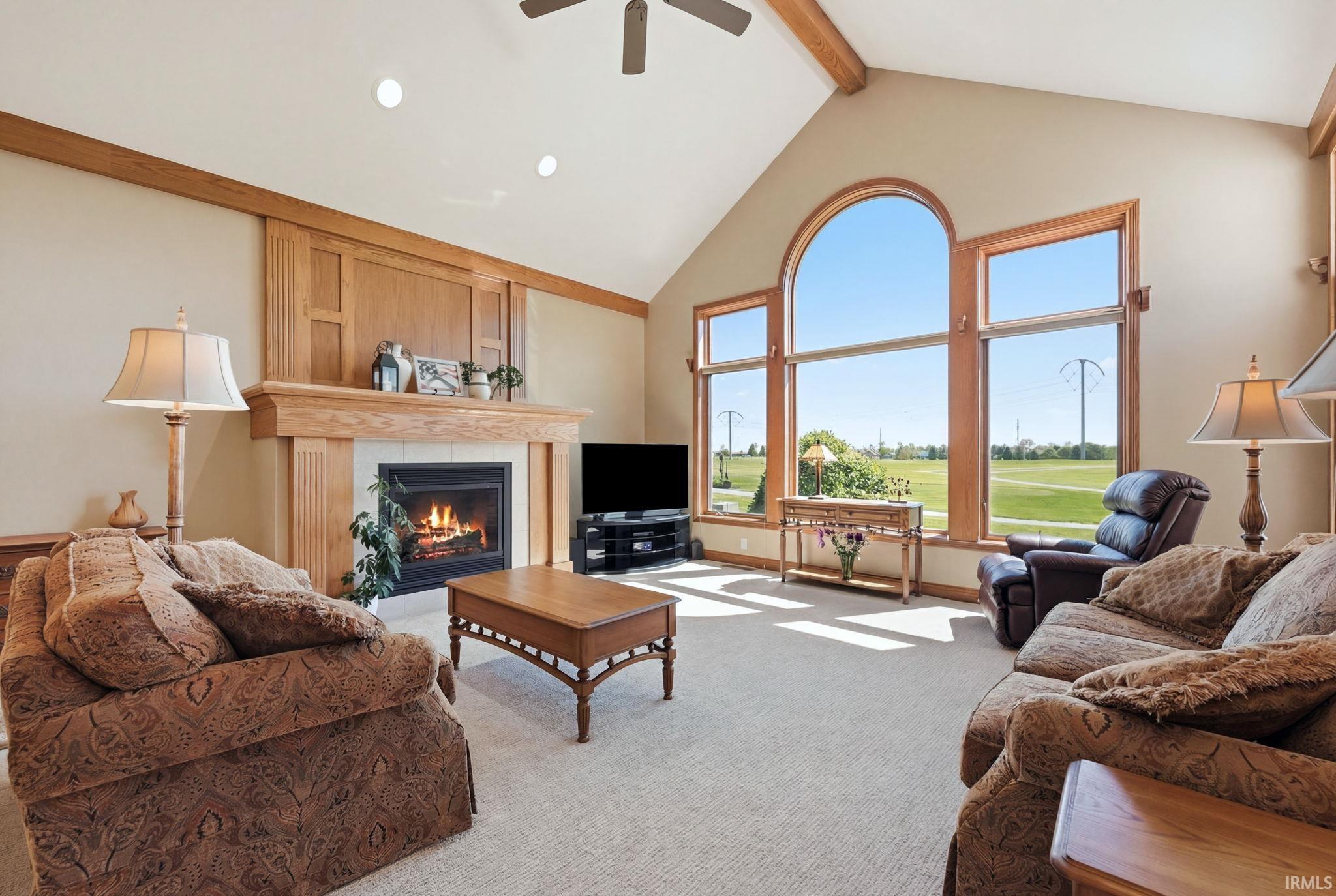 Vaulted ceiling living room featuring a floor-to-ceiling window arrangement with an arched top, a wood-finish fireplace mantel with a hearth, exposed ceiling beams, and recessed lighting