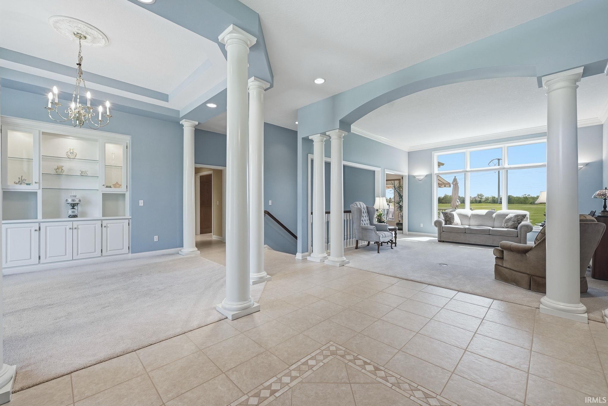 Spacious interior featuring tile flooring with decorative inlay, architectural columns, a built-in display cabinet with glass doors, a chandelier, and large windows
