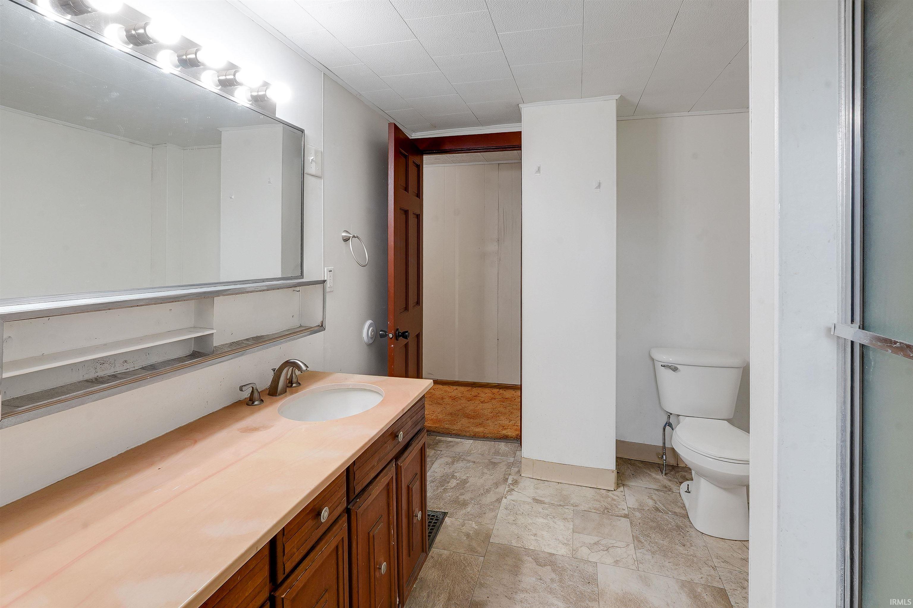 Bathroom featuring a brown wood-finish vanity with an integrated sink and brushed nickel faucet, a large mirror with overhead strip lighting, tiled flooring, and a white toilet