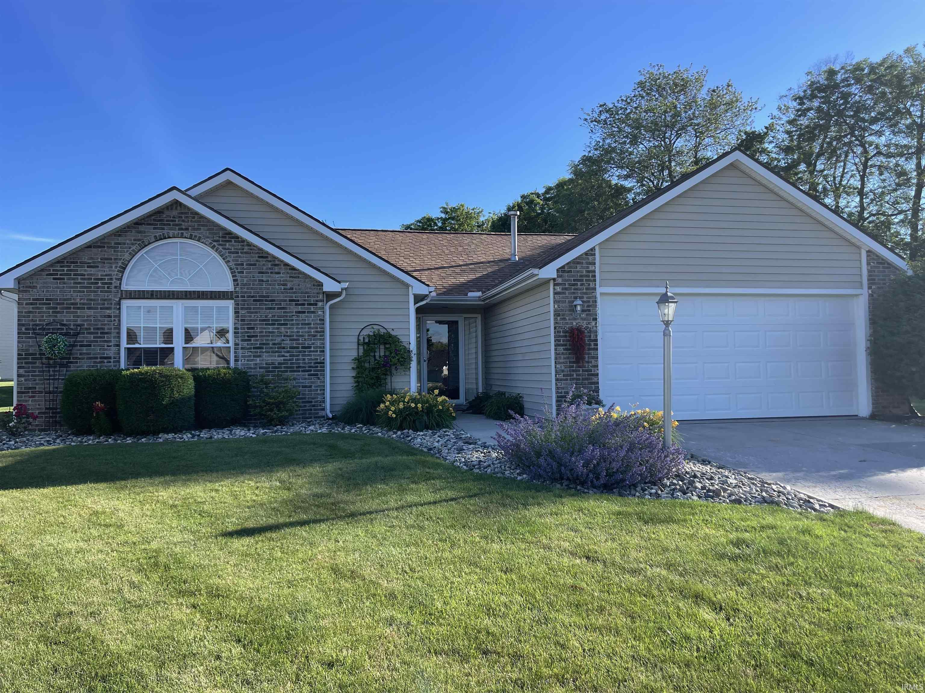 Single-story residence featuring a brick facade with an arched window, light-colored siding, an attached two-car garage, manicured lawn, and decorative landscaping