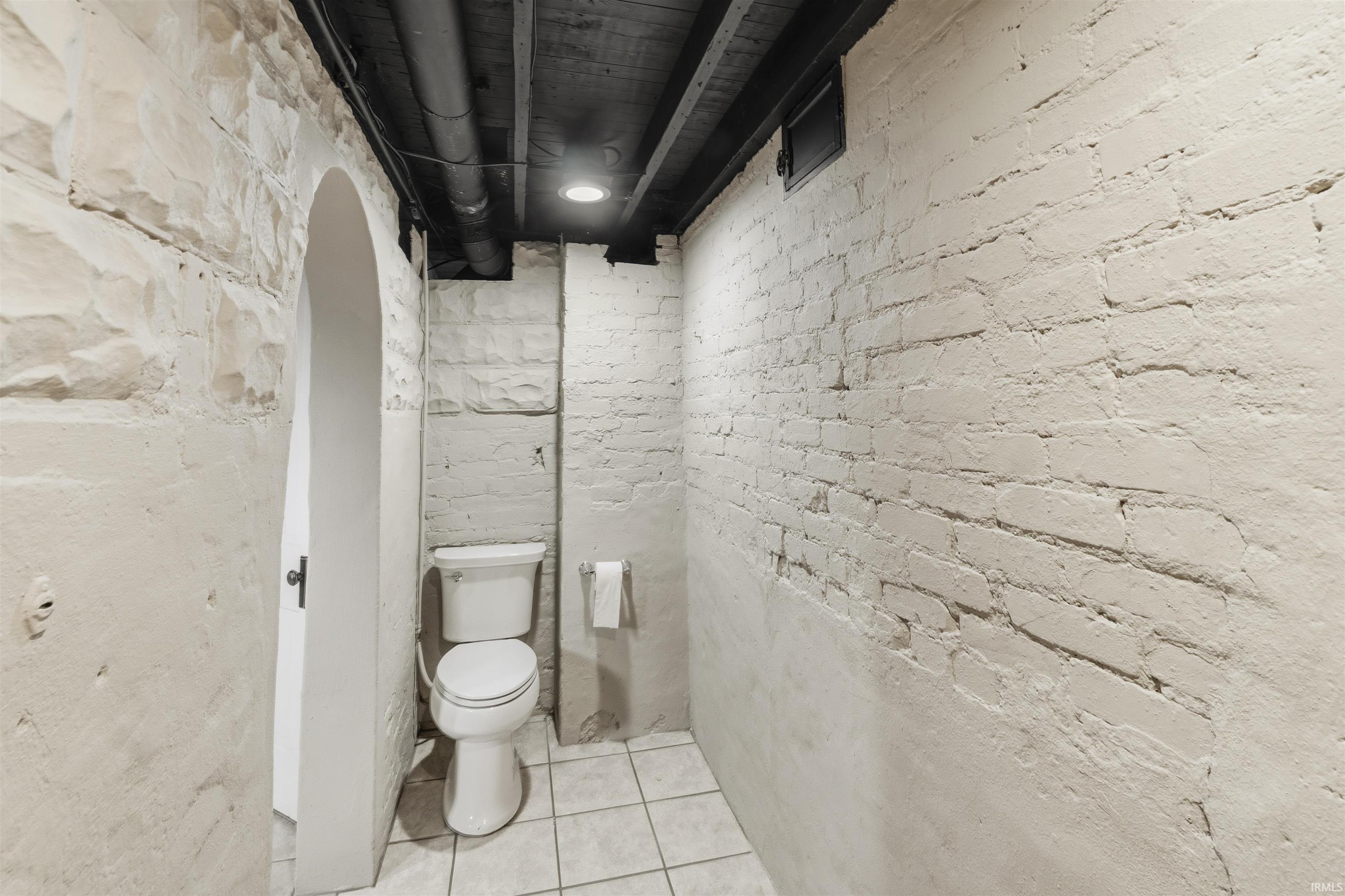 Compact bathroom featuring a white porcelain toilet, white square floor tiles, white-painted exposed brick walls, and a recessed ceiling light