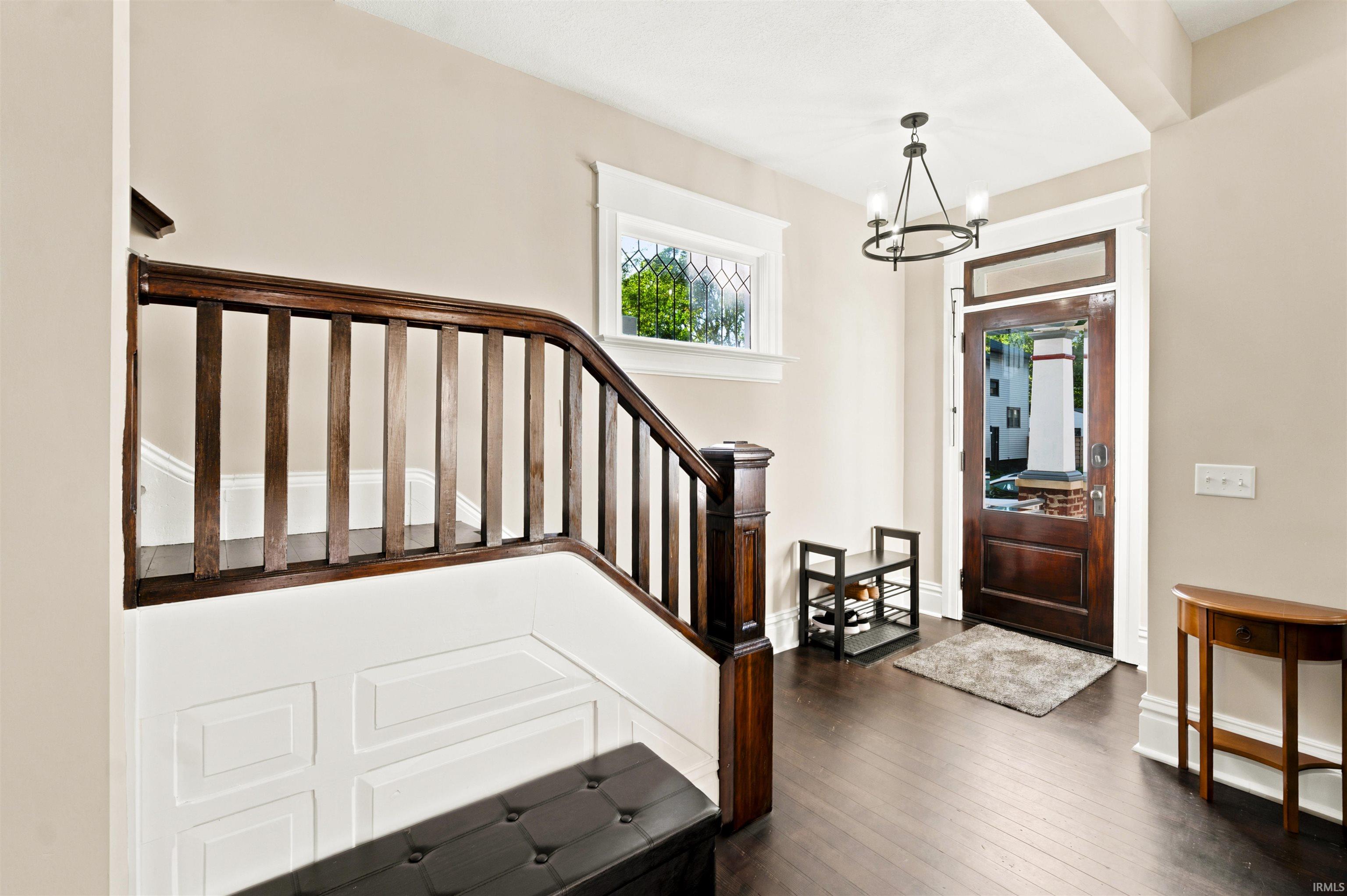 Entryway featuring dark wood-finish flooring, a dark wood staircase with paneling, a dark wood entry door with glass paneling, a transom window, and a chandelier with a circular frame