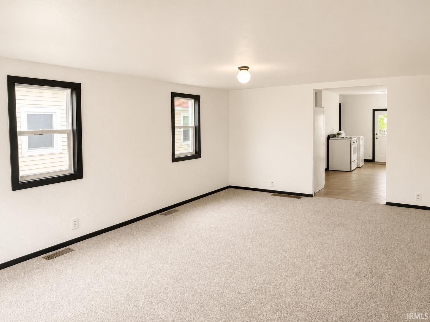 Open concept living space featuring neutral wall tones, light-colored carpeting, two windows with black trim, and a ceiling-mounted light fixture