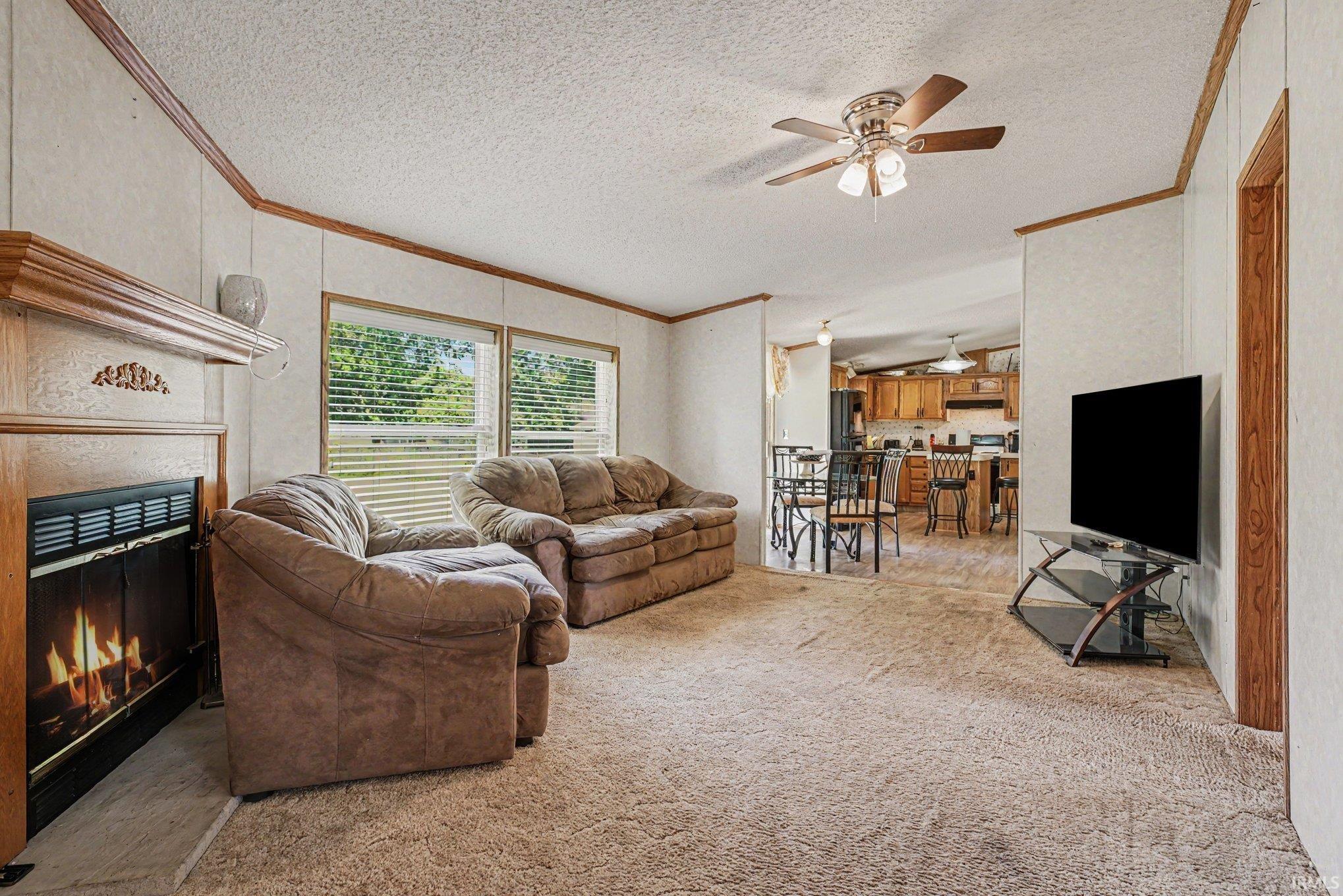 Living area featuring a gas fireplace with a wood mantle, carpet flooring, a ceiling fan, and multiple windows