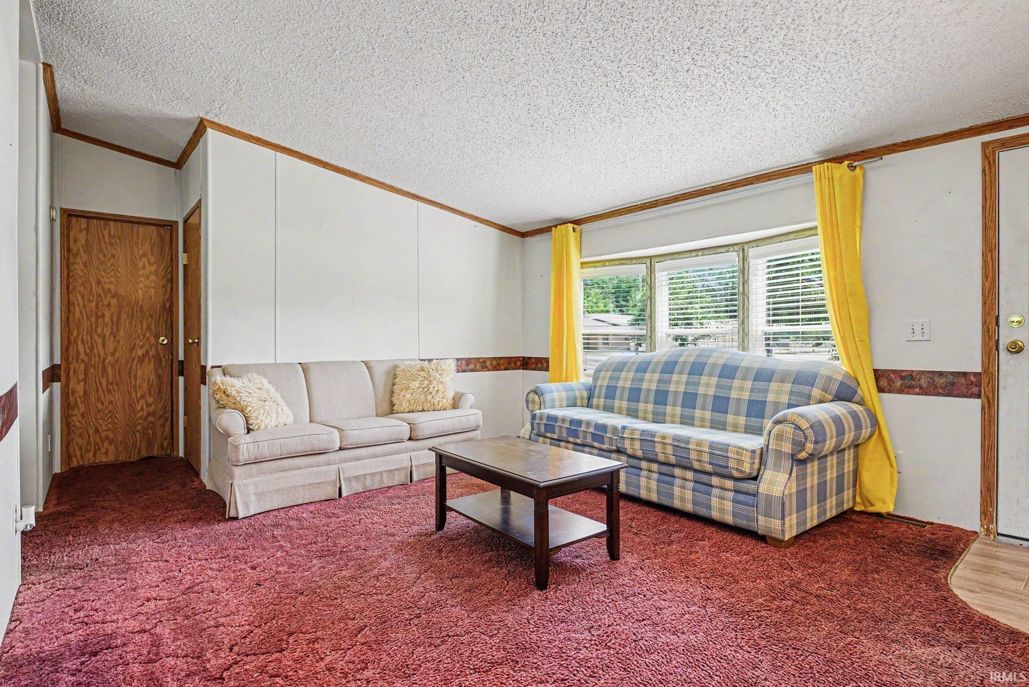 Living area featuring red textured carpeting, wood-paneled walls with white sections, a bay window with horizontal blinds, and a wood-finish entry door with coordinating trim