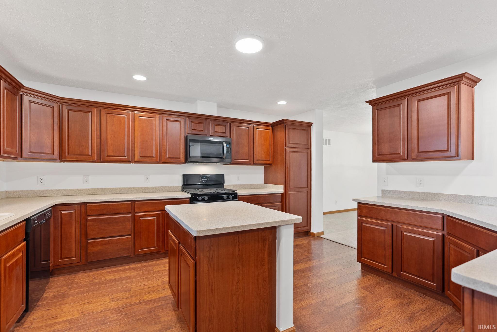 Spacious kitchen featuring extensive wood cabinetry, light-toned countertops, a central island, integrated black appliances, and wood-finish flooring