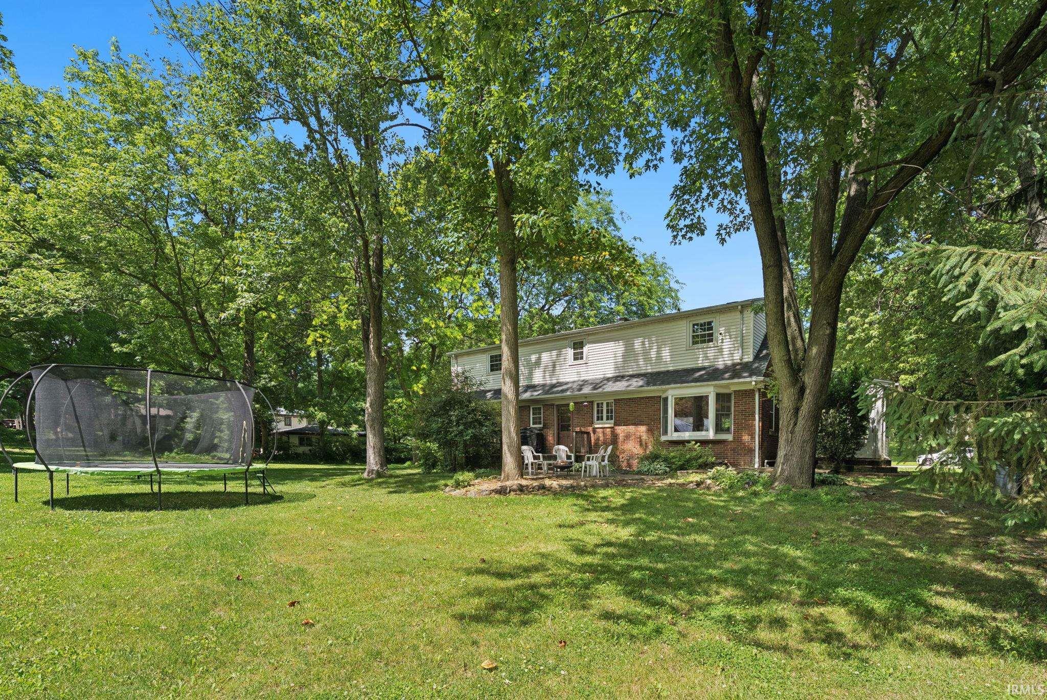 Expansive green lawn with mature trees and a two-story residence featuring a brick facade on the lower level and light-colored siding on the upper level