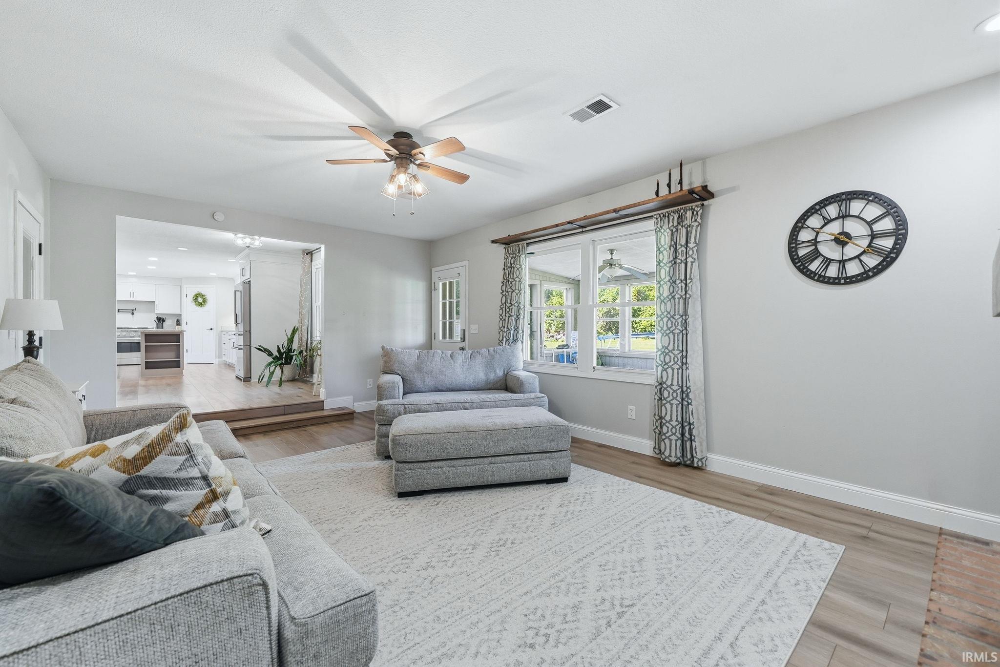 Spacious living area featuring light wood-finish flooring, a ceiling fan, and large windows with patterned curtains