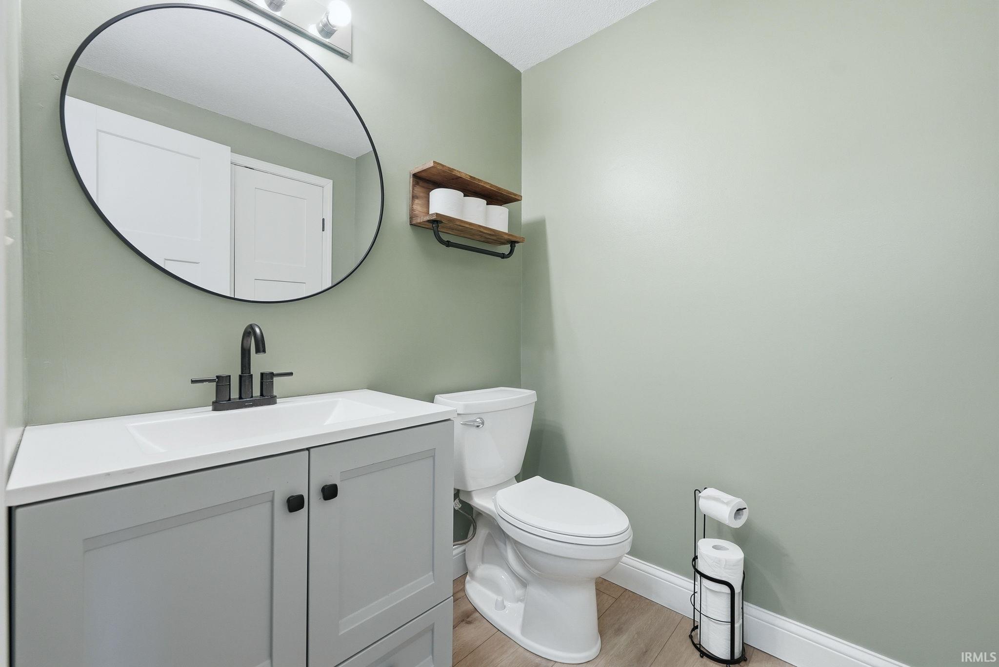 Powder room featuring a gray shaker-style vanity with a white countertop and integrated sink, a black-framed oval mirror, and light wood-finish flooring
