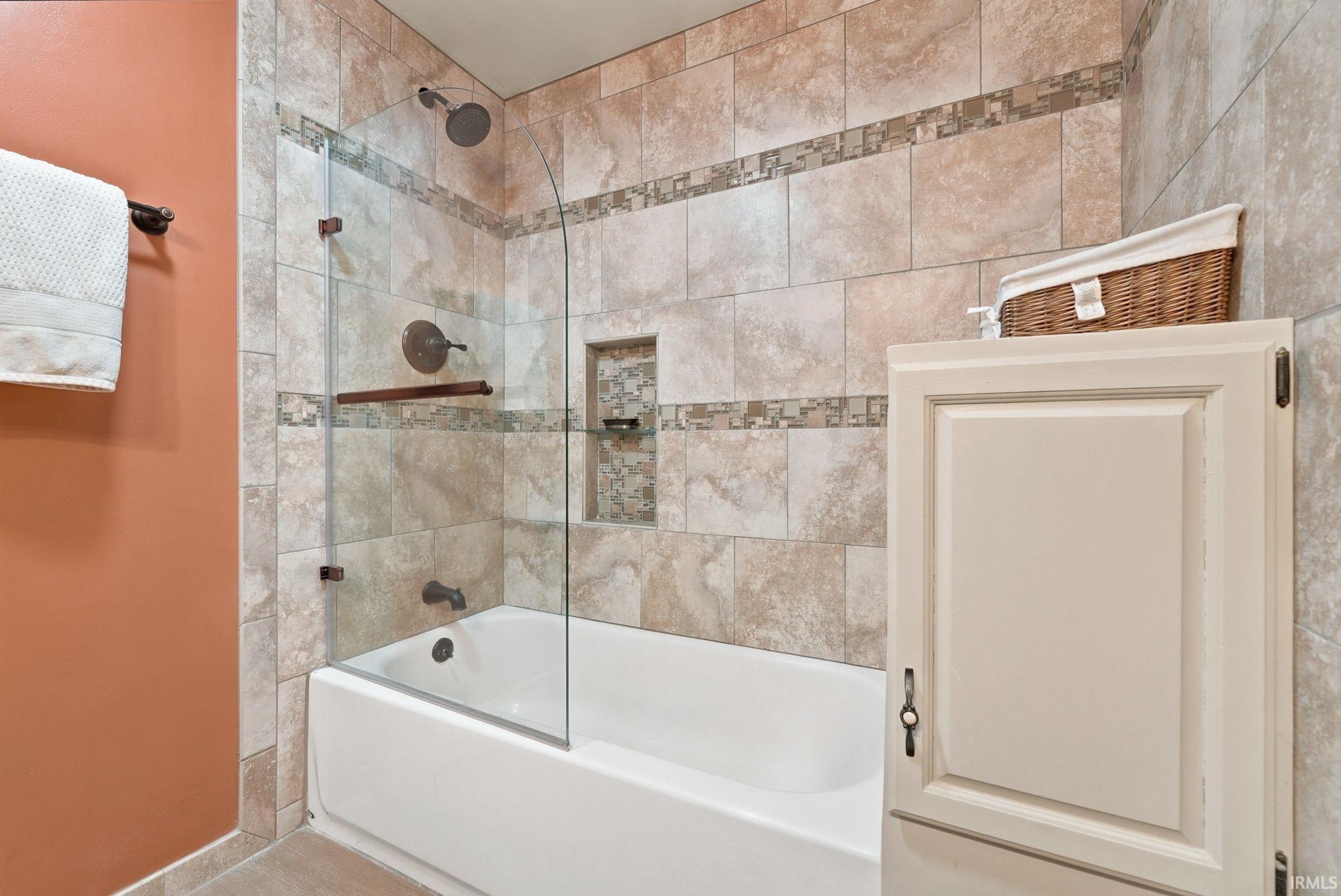 Tub and shower combination featuring extensive neutral-toned wall tiling, a decorative mosaic tile accent, a built-in shower niche with shelving, and a clear glass shower screen