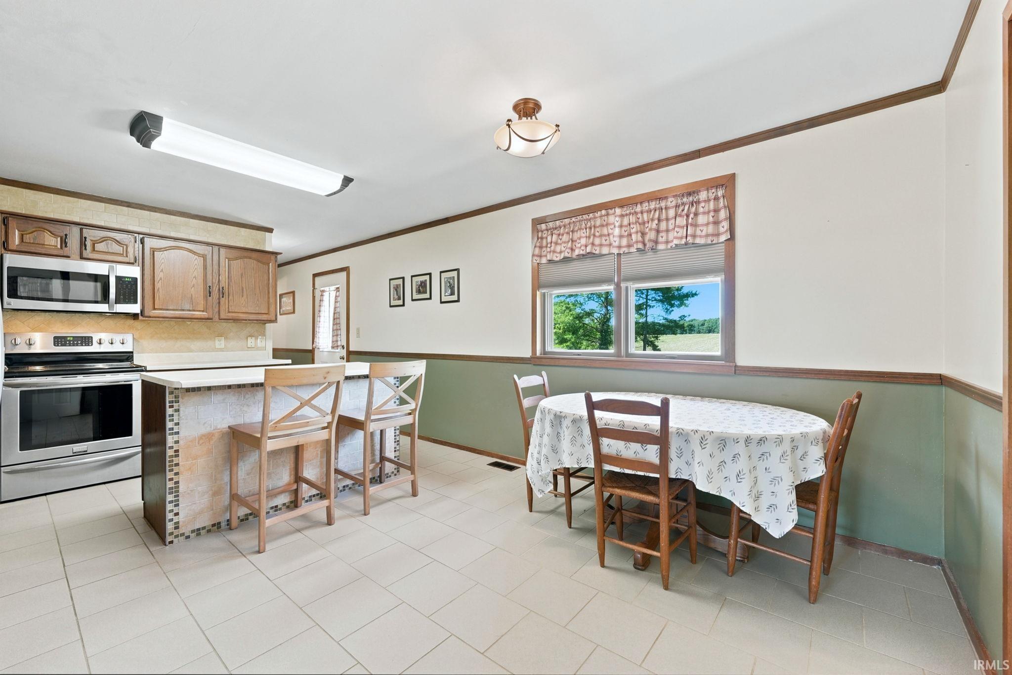 Open-concept kitchen and dining area featuring light-toned tile flooring, an island with mosaic tile accents, stainless steel appliances, and a window framing a rural landscape