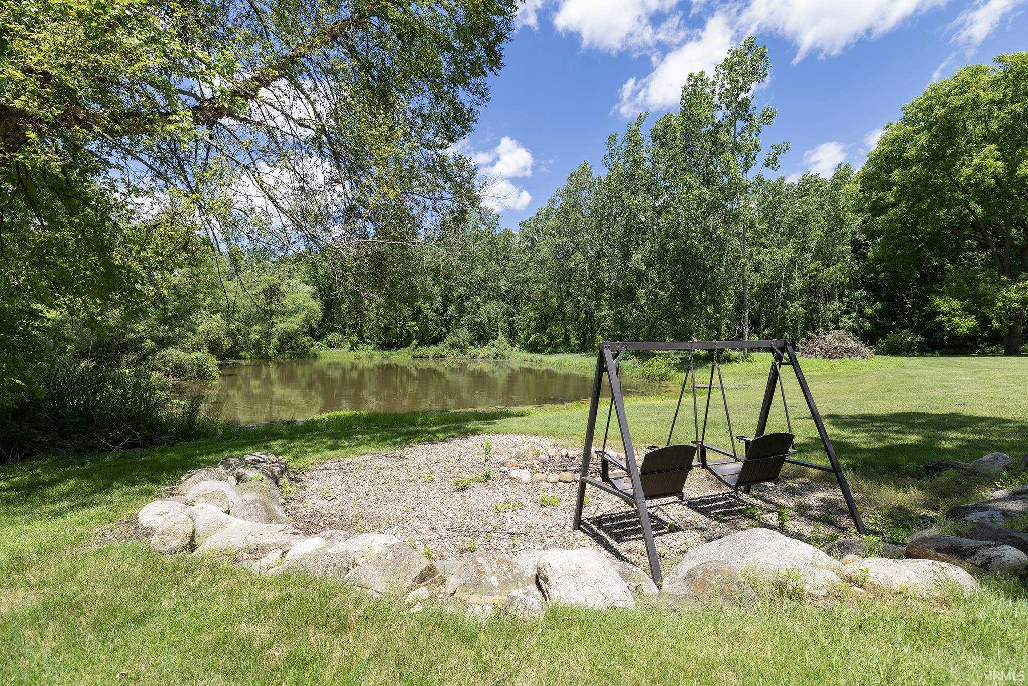 Waterside fire pit area with stone surround, gravel base, and a metal porch swing, surrounded by a expansive lawn and mature trees