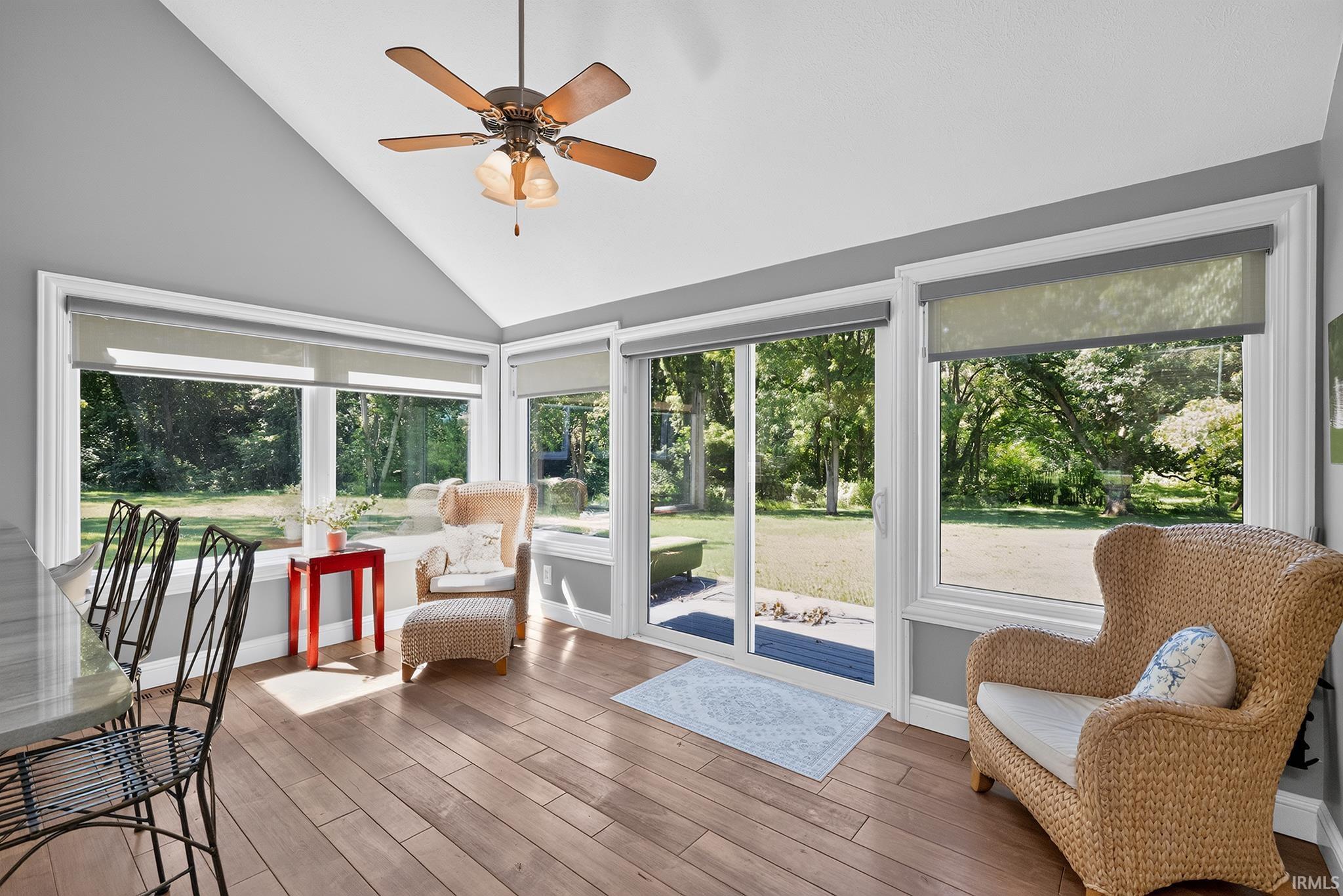 Sunroom featuring vaulted ceilings, wood-finish flooring, a ceiling fan, multiple windows with roller shades, and a glass sliding door