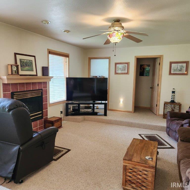 Living area featuring a tiled fireplace with a wood mantle, recessed lighting, a ceiling fan with an ornate light fixture, wood-trimmed windows, and light-colored carpeting