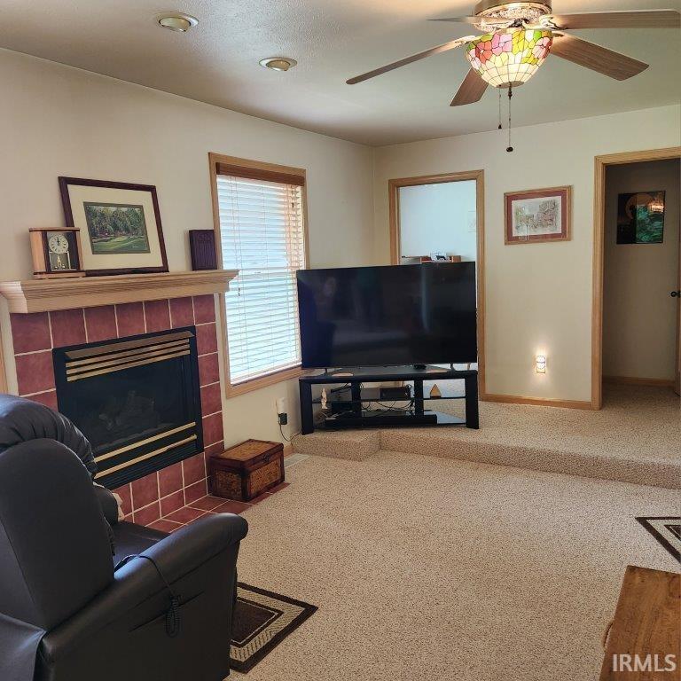Carpeted living space featuring a tiled fireplace with a wood mantle, a window with horizontal blinds, and a decorative ceiling fan with a stained-glass light fixture