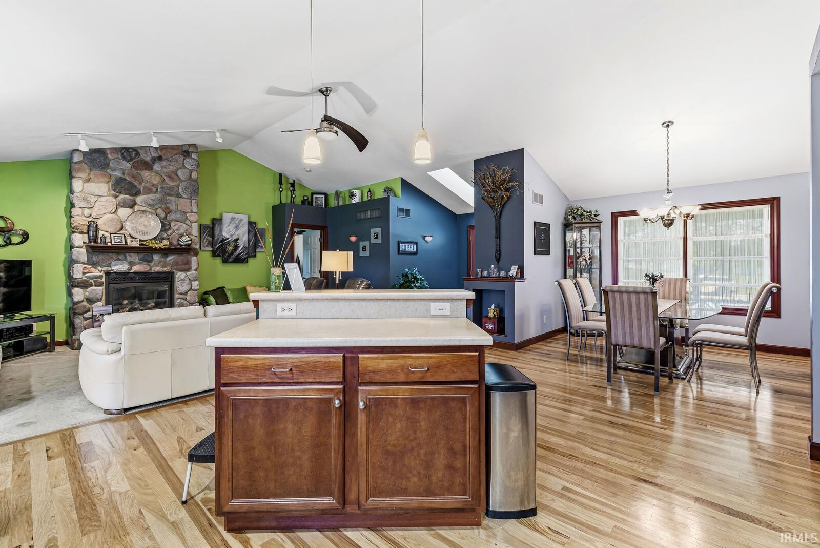 Open-concept living space featuring wood-finish flooring, a stone fireplace, vaulted ceilings with ceiling fans, a kitchen island with storage, and a dining area with a chandelier