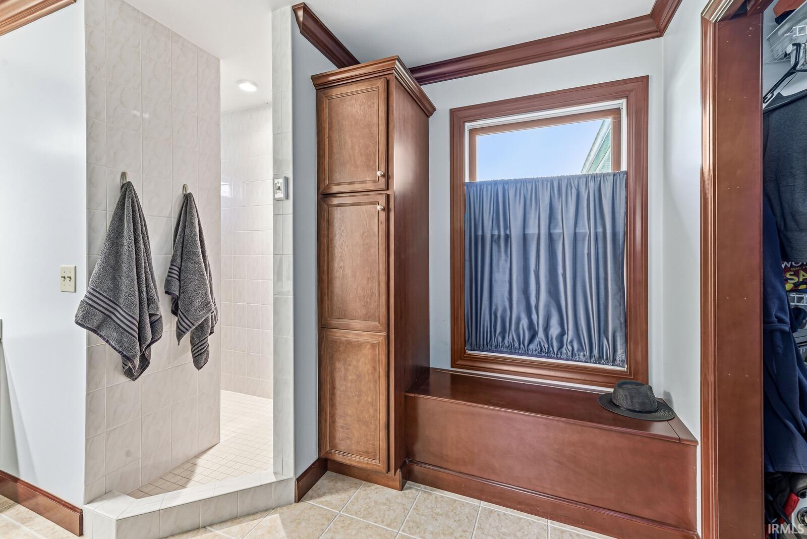 Bathroom area featuring a tiled shower enclosure, wood-finish built-in cabinetry, a window with wood-finish trim, a built-in storage bench, and ceramic tile flooring