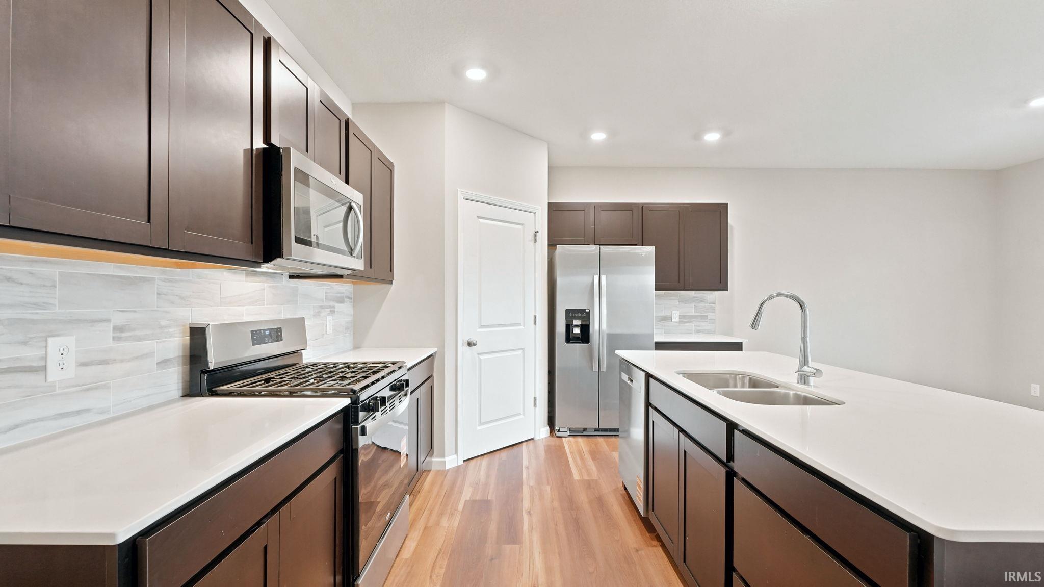 Modern kitchen featuring dark wood cabinetry, stainless steel appliances, a white tile backsplash, light countertops, and wood-finish flooring