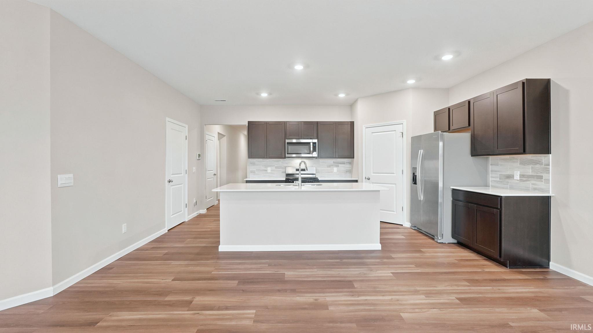 Open-concept kitchen featuring an island with a white countertop, dark wood-finish cabinetry, stainless steel appliances, and wood-finish flooring