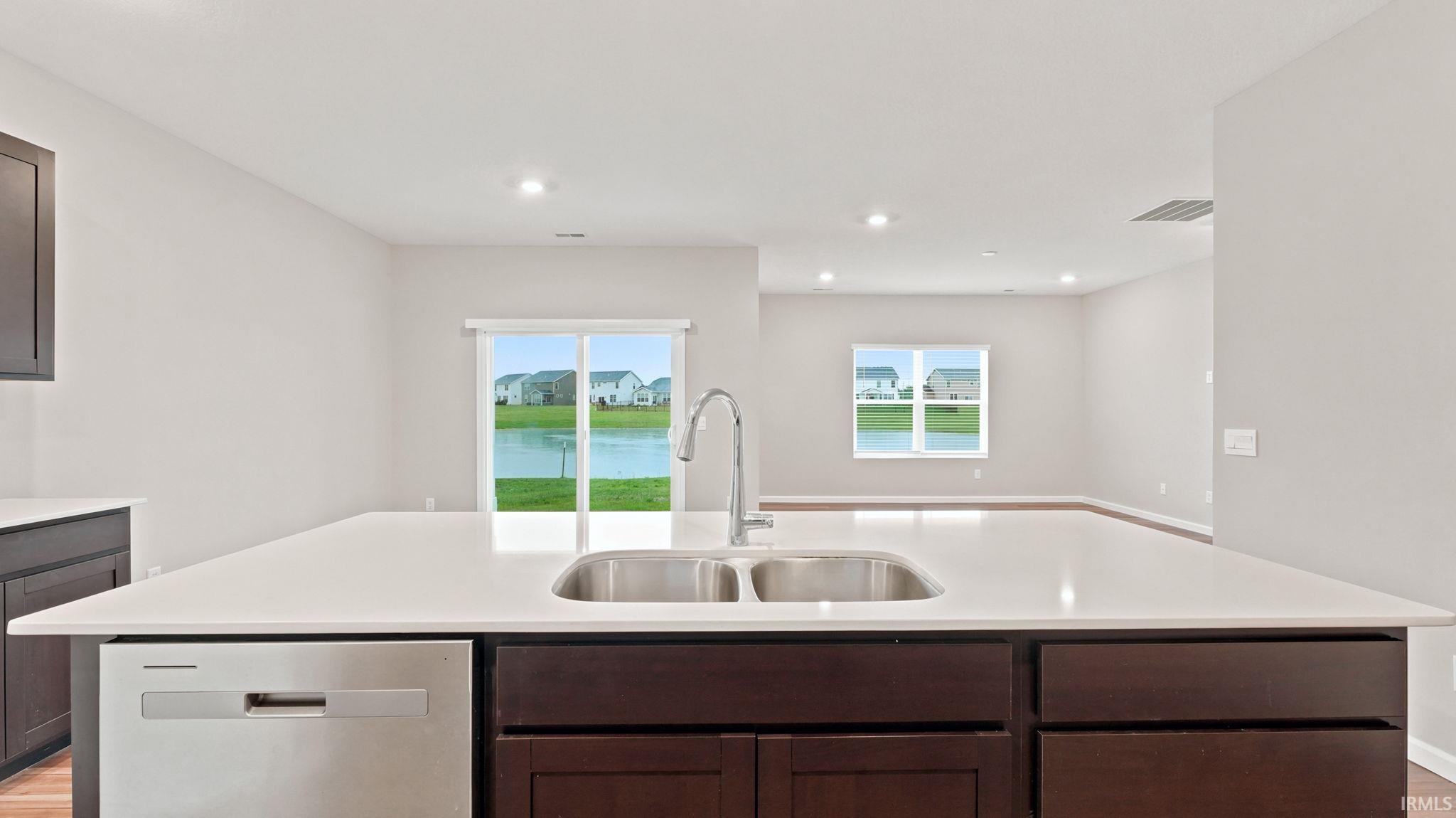 Kitchen island featuring a double basin stainless steel sink with a high-arc faucet, integrated stainless steel dishwasher, light-toned countertop, and dark wood-finish cabinetry