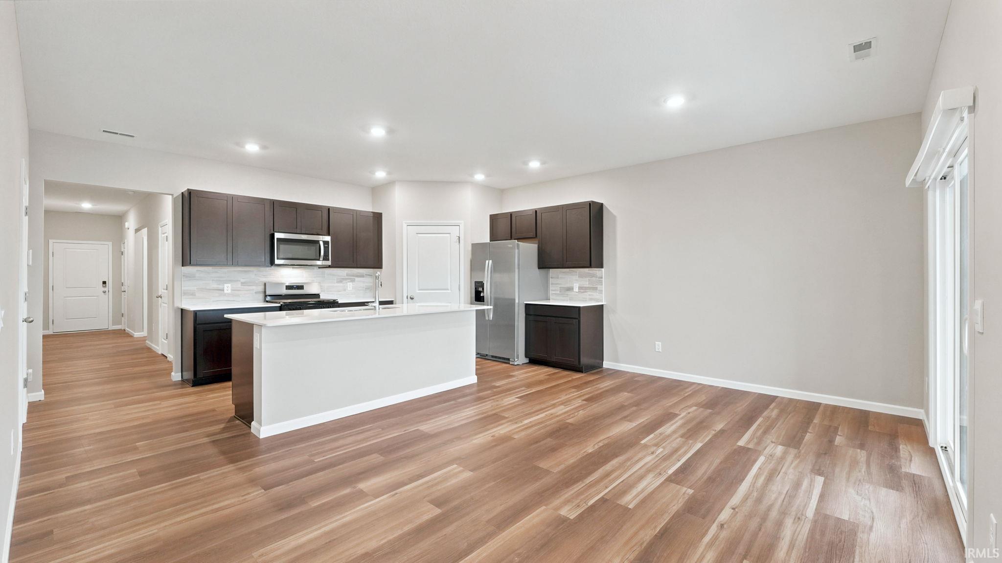Open-concept kitchen and living area featuring wood-finish flooring, dark wood cabinetry, stainless steel appliances, and a central island with a breakfast bar