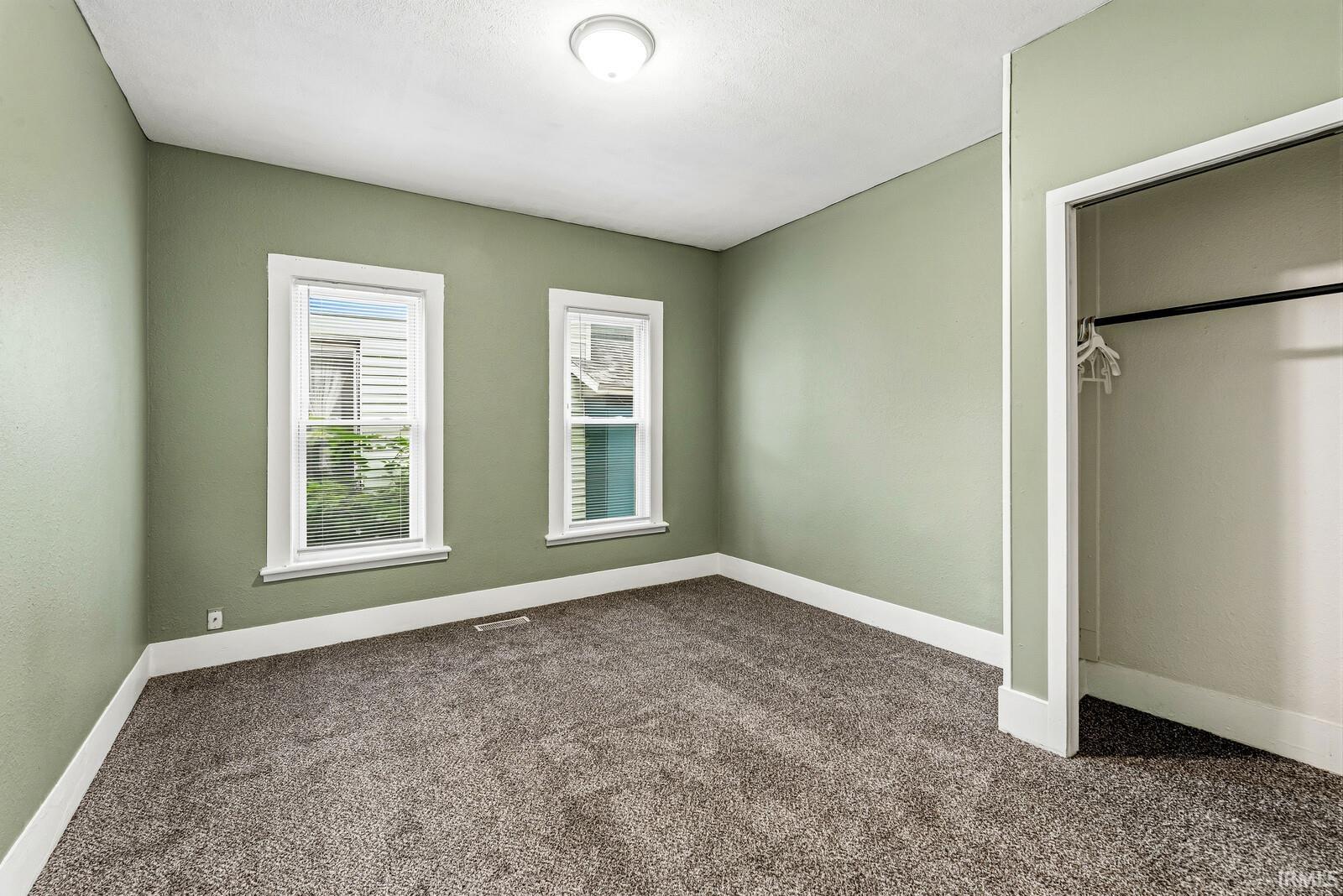 Carpeted room with two white-trimmed windows, a flush-mount ceiling light, and a closet featuring a hanging rod