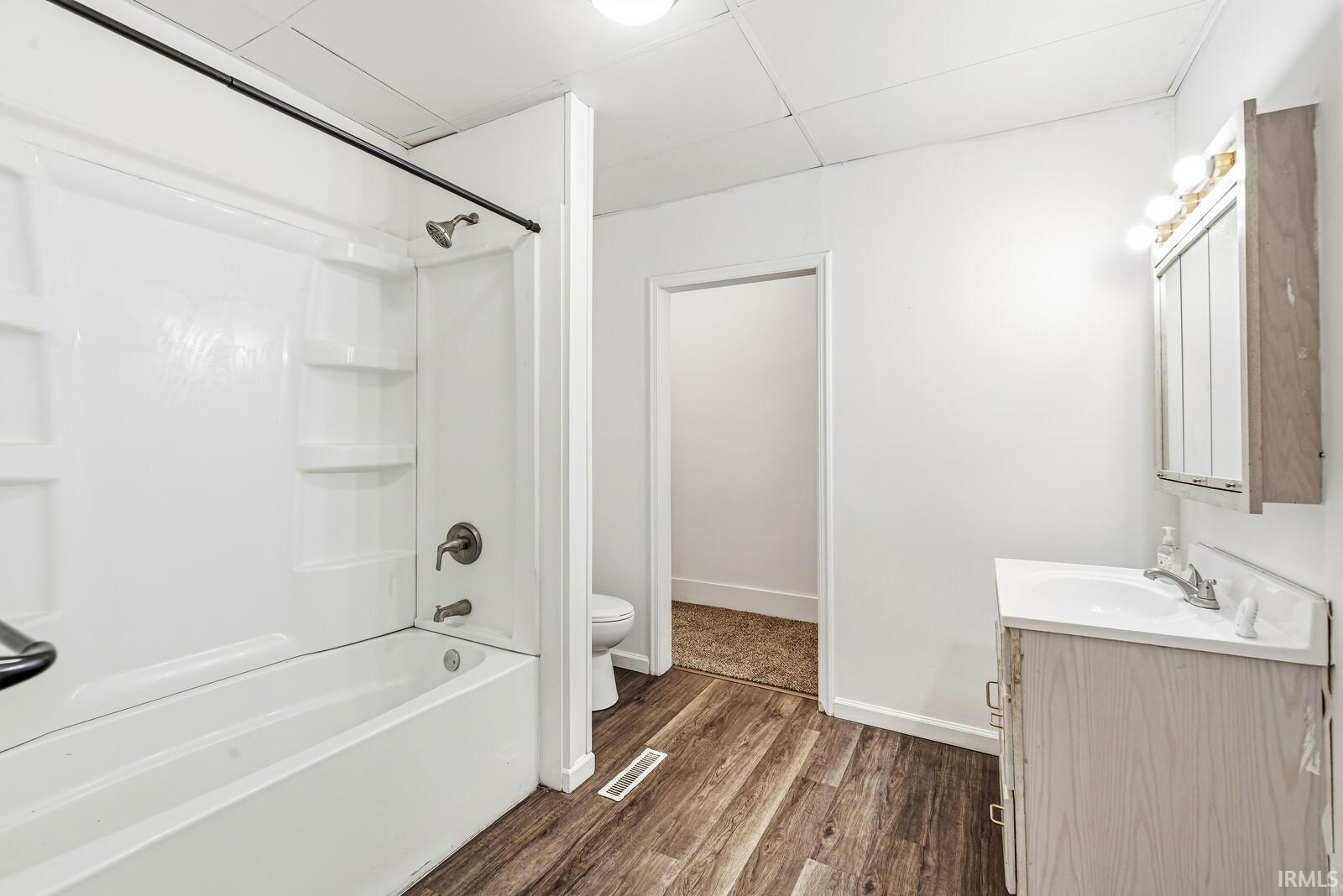 Bathroom featuring wood-finish flooring, a white vanity with a single sink, an overhead wall cabinet, and a bathtub with an integrated shower and white surround