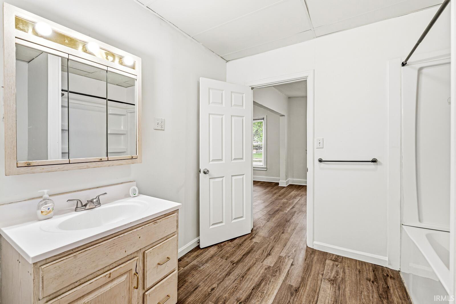 Bathroom featuring a wood-finish vanity with a single basin sink and chrome faucet, a mirrored medicine cabinet with integrated lighting, a white bathtub with a shower rod, and wood-finish flooring