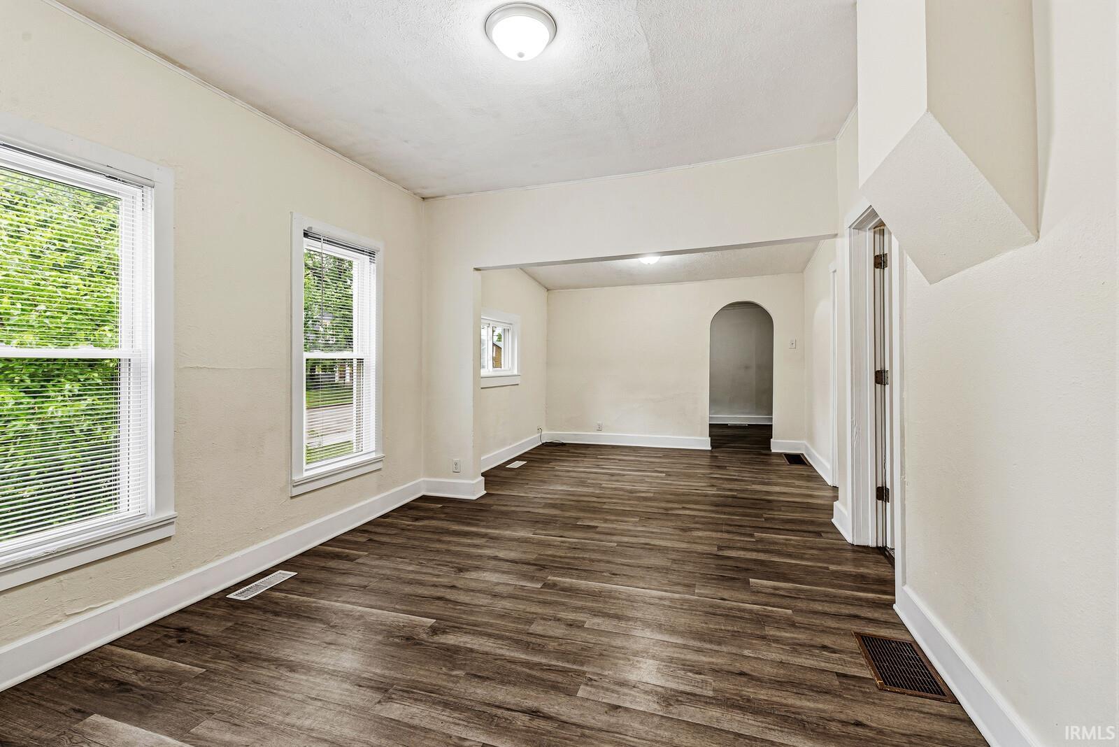 Spacious room featuring wood-finish flooring, two large windows with blinds, a ceiling-mounted light fixture, and an arched doorway