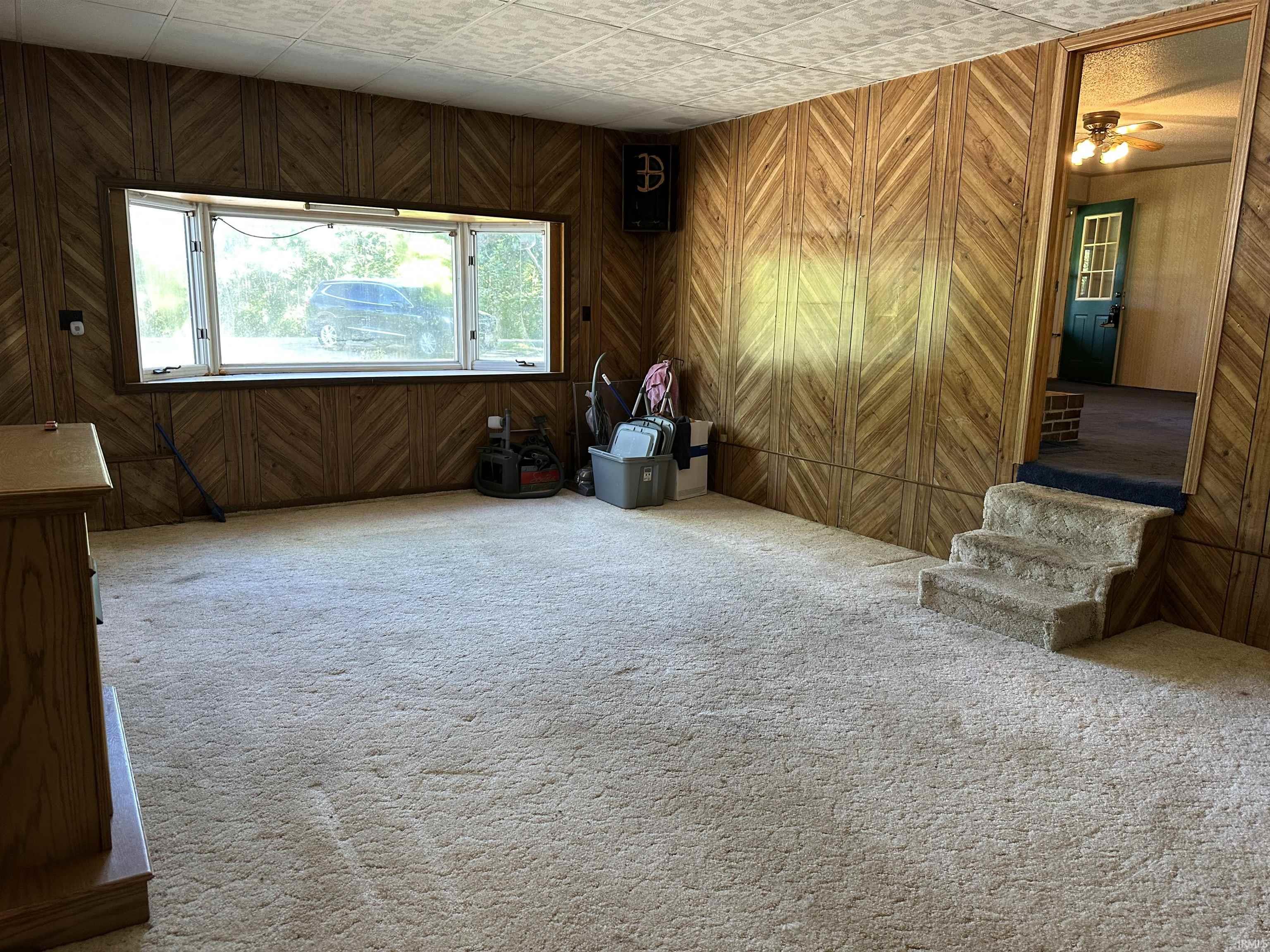 Spacious room featuring light-colored carpet flooring, a large bay window, wood-finish wall paneling, and a three-step carpeted staircase leading to an arched doorway