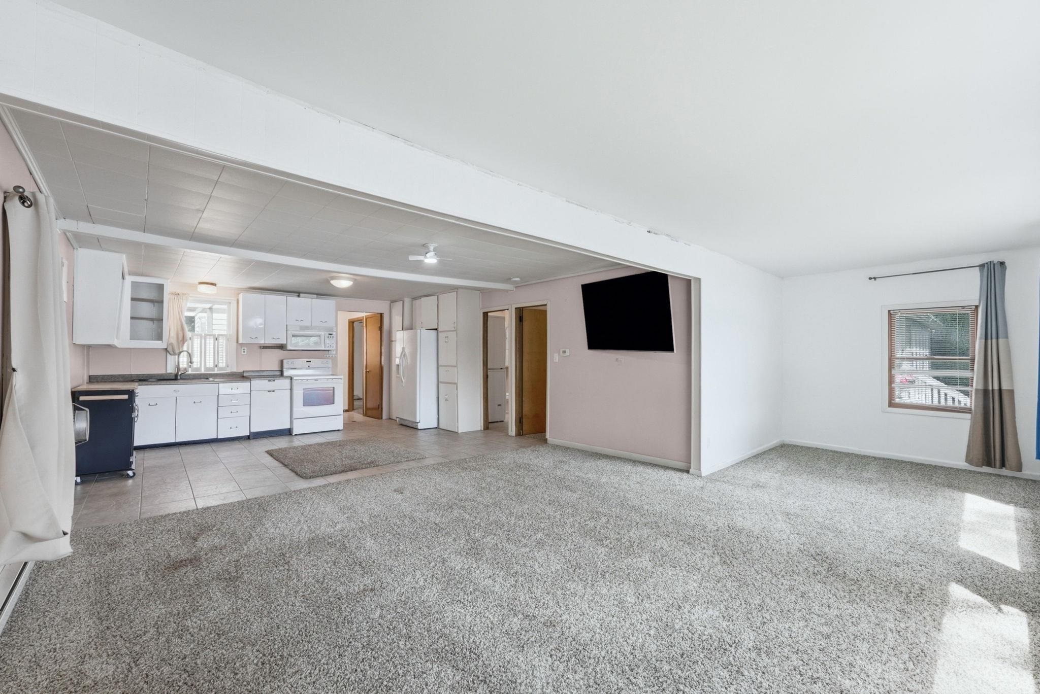 Open-concept living area with light-colored carpeting, a wall-mounted display, and a window with a curtain rod