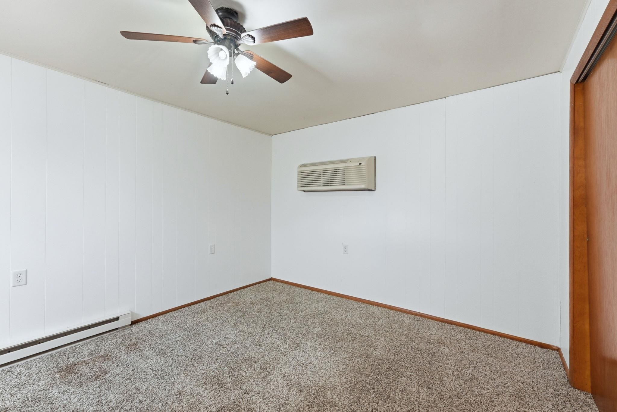 Carpeted room featuring a ceiling fan with integrated lighting, white panel walls, a wall-mounted air conditioning unit, and a built-in closet with a wood-finish door