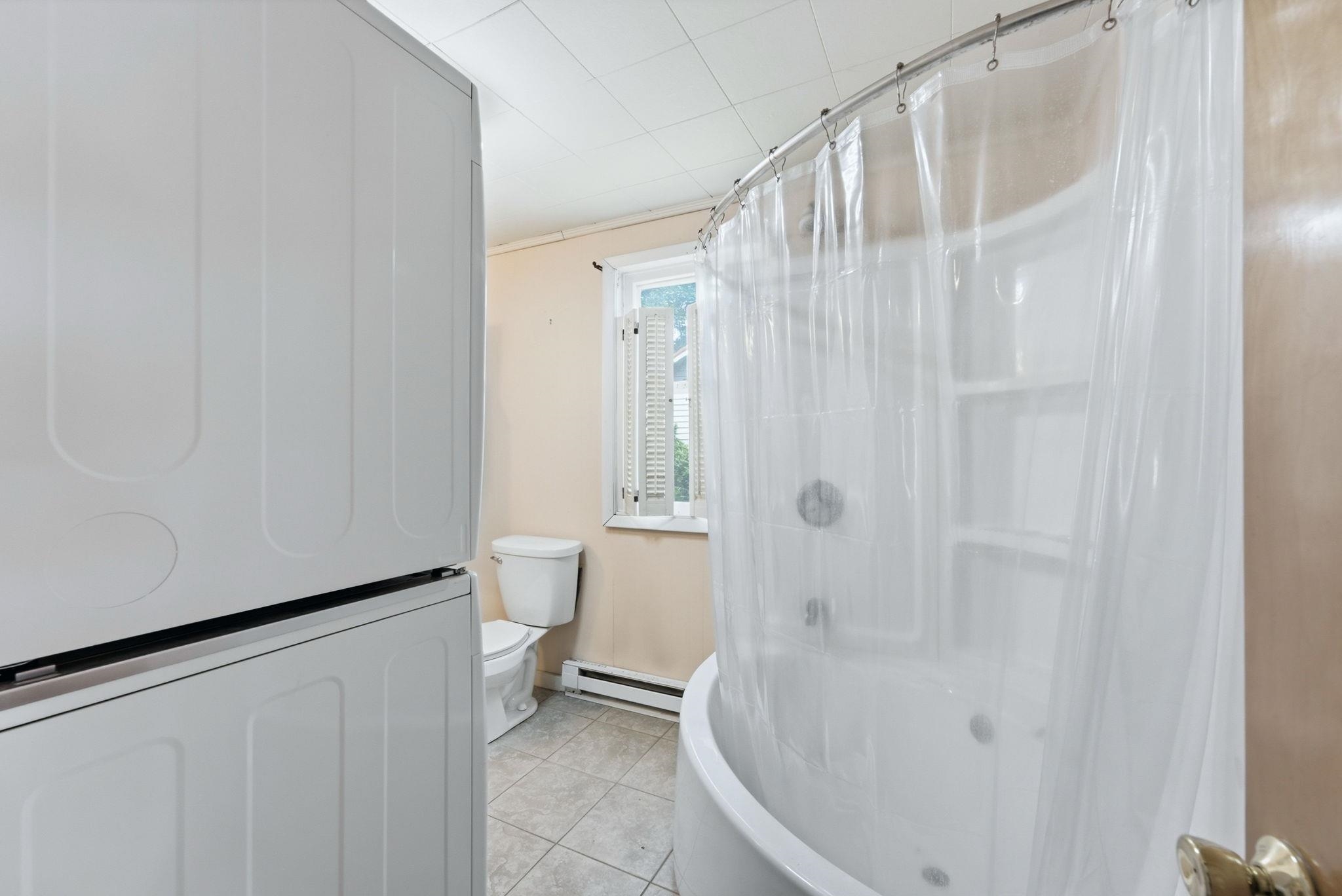 Bathroom featuring a curved shower-tub combination, a toilet, a window with louvered shutters, and tiled flooring