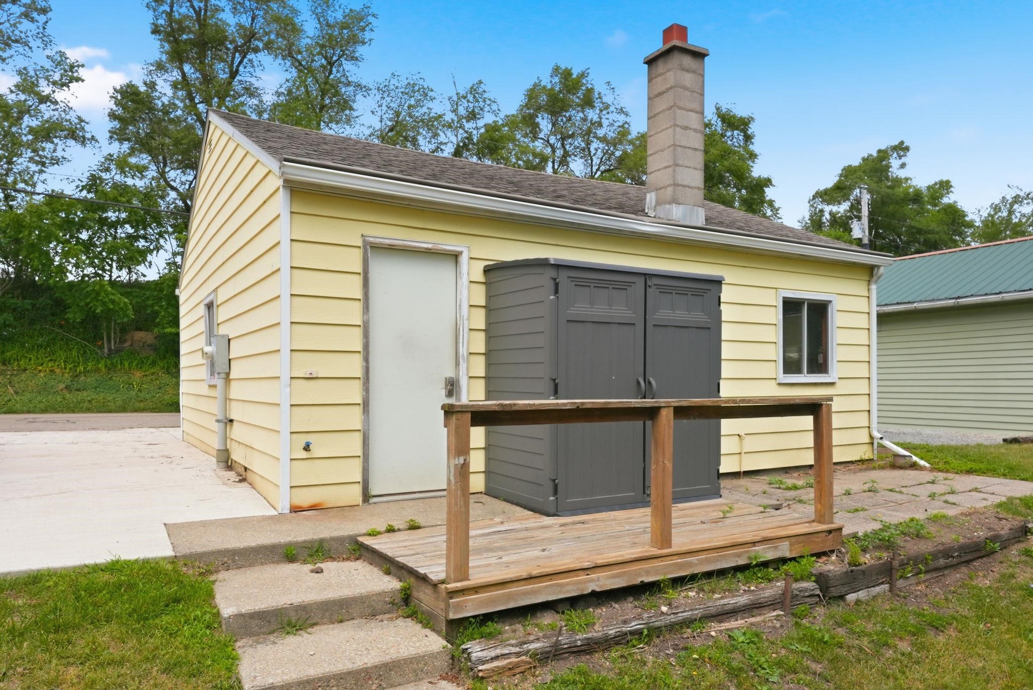 Yellow siding structure featuring a wood deck with railing, concrete patio, and a stone chimney