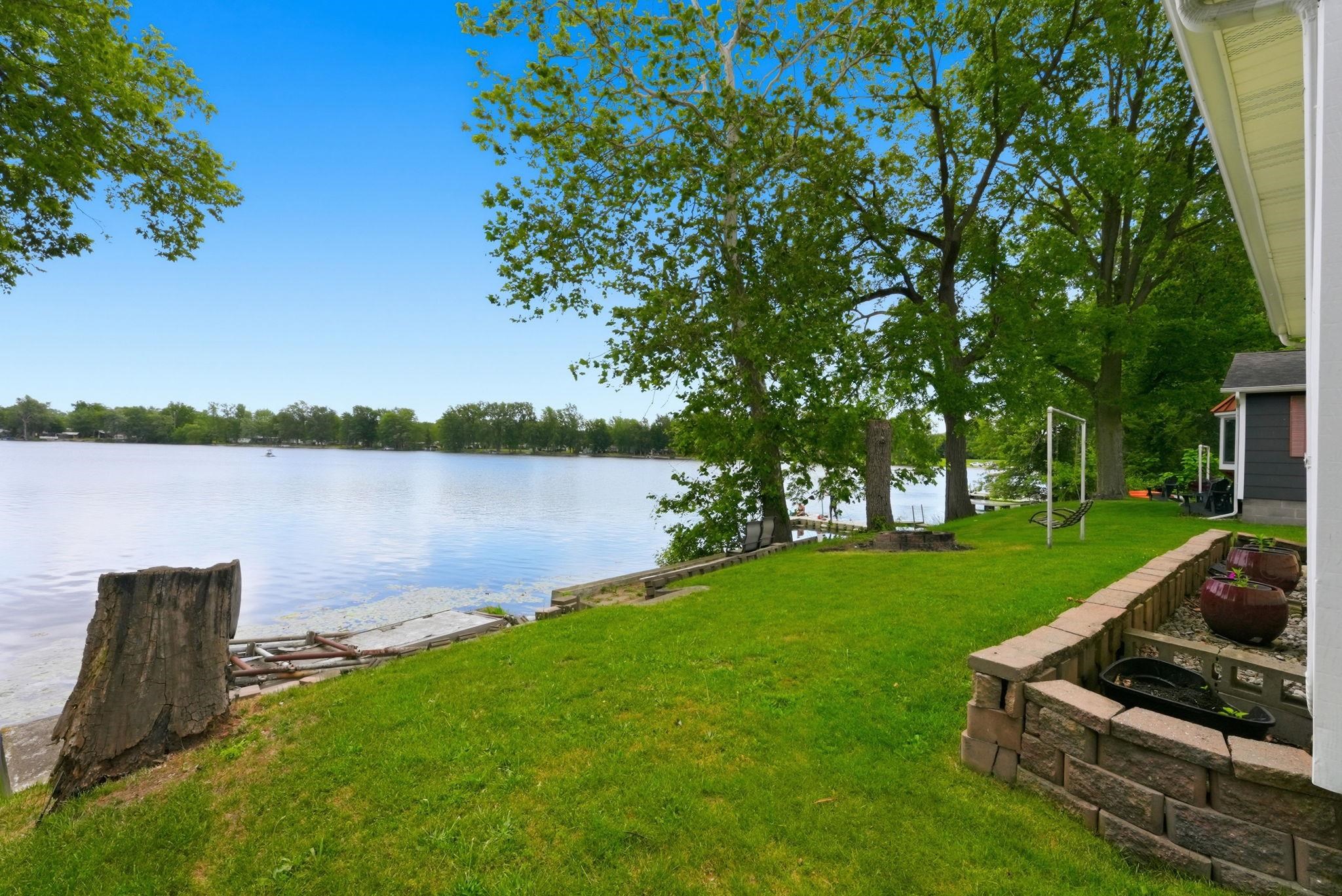 Lakeside property featuring a spacious grassy area, mature trees, and a stone block retaining wall with integrated planters