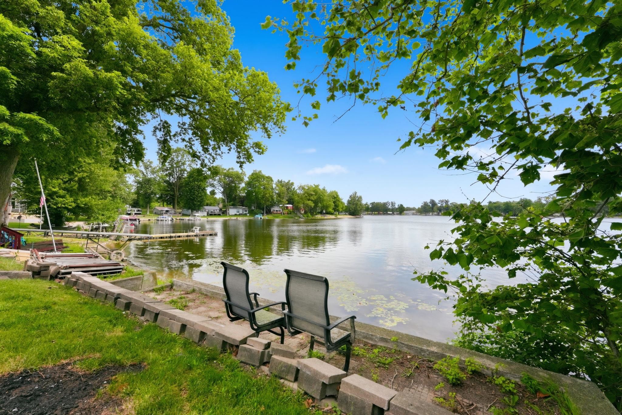 Expansive waterfront setting featuring a tranquil lake, mature trees providing natural shade, a concrete retaining wall, and a wooden dock structure