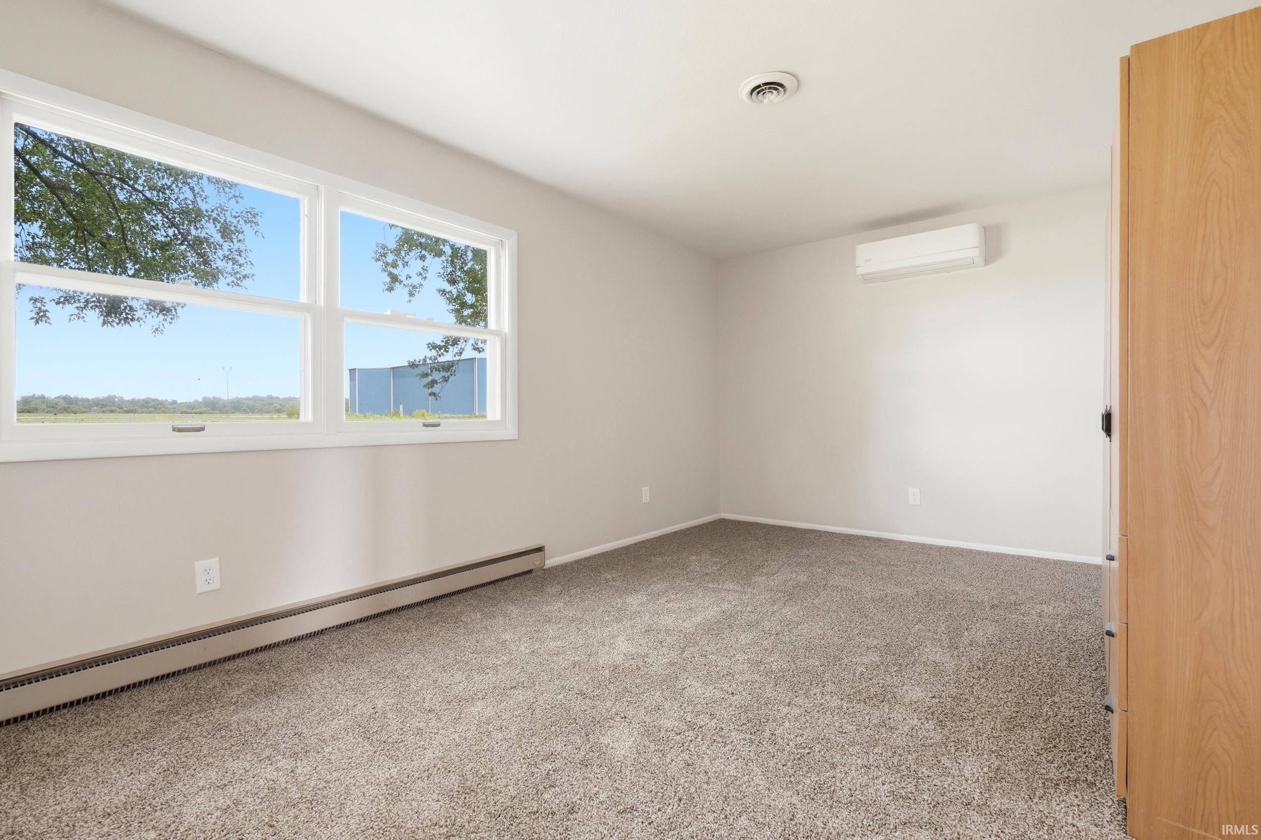 Spacious room featuring neutral carpeting, a large window, light-colored walls, baseboard heating, and an installed wall-mounted air conditioning unit