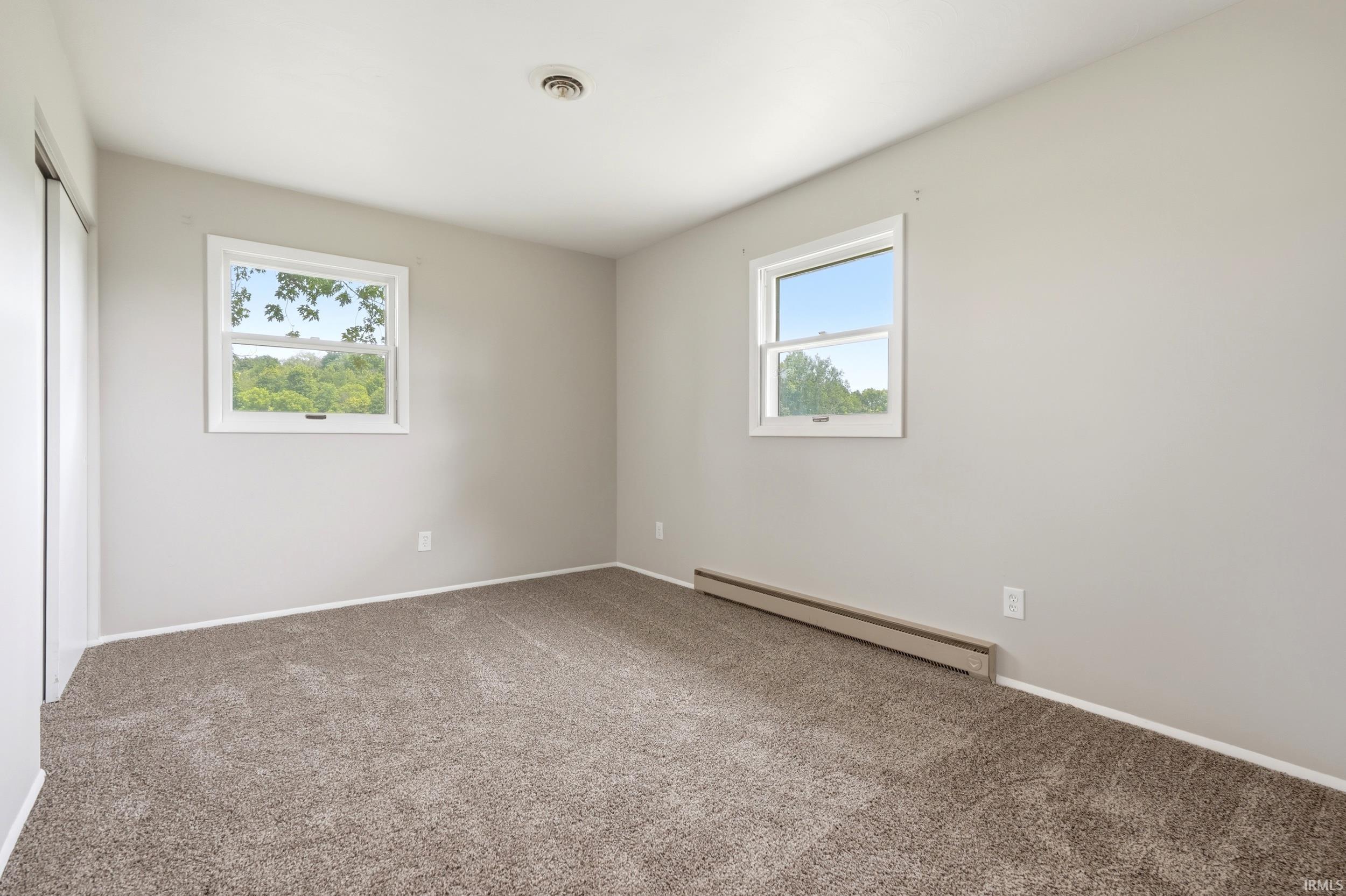 Spacious room featuring neutral paint, plush carpeting, two windows, and baseboard heating