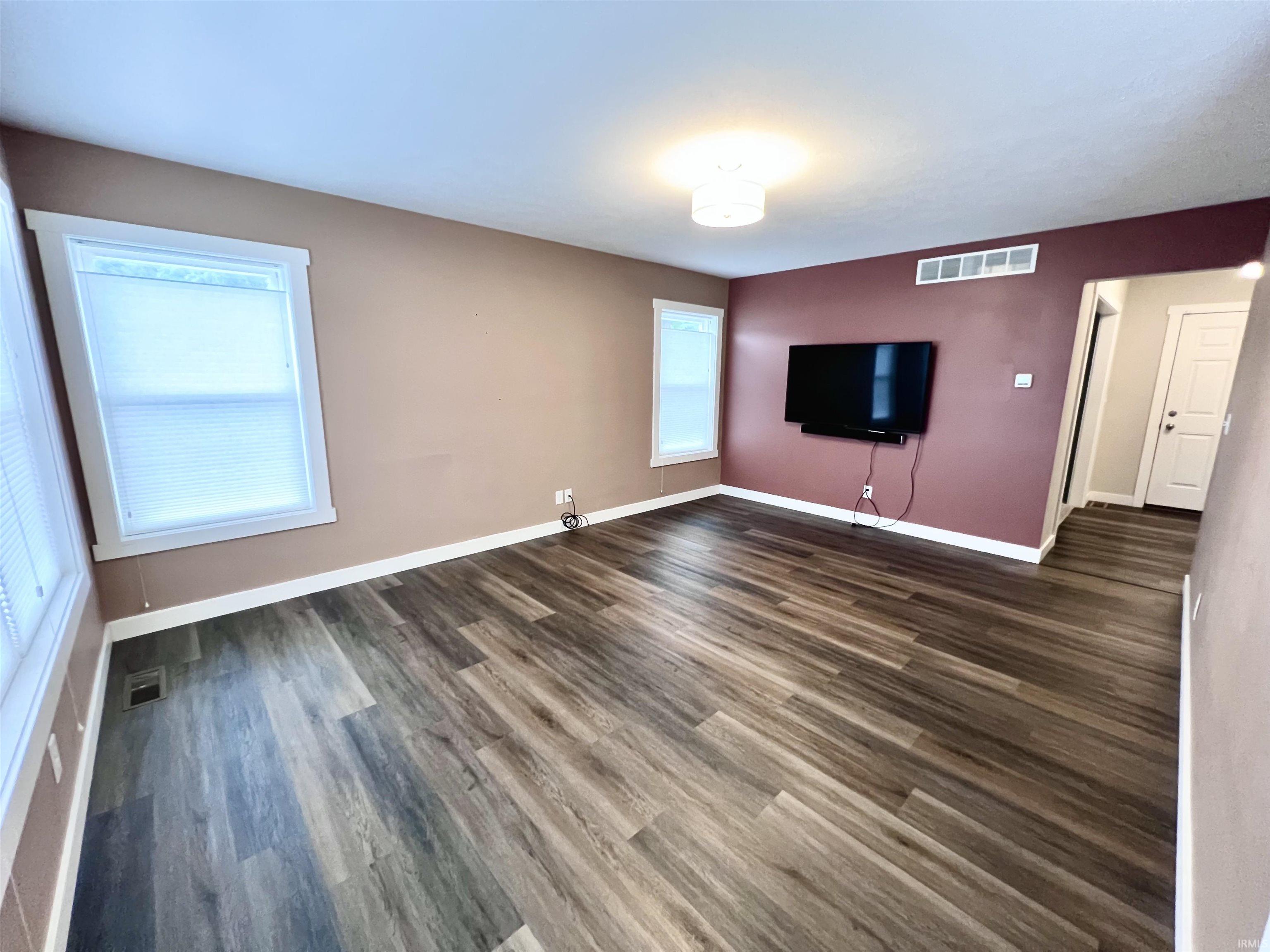 Spacious open floor plan featuring wood-finish flooring throughout, neutral wall palette with accent burgundy wall, large window with natural light, wall-mounted flat-screen television, and recessed ceiling lighting