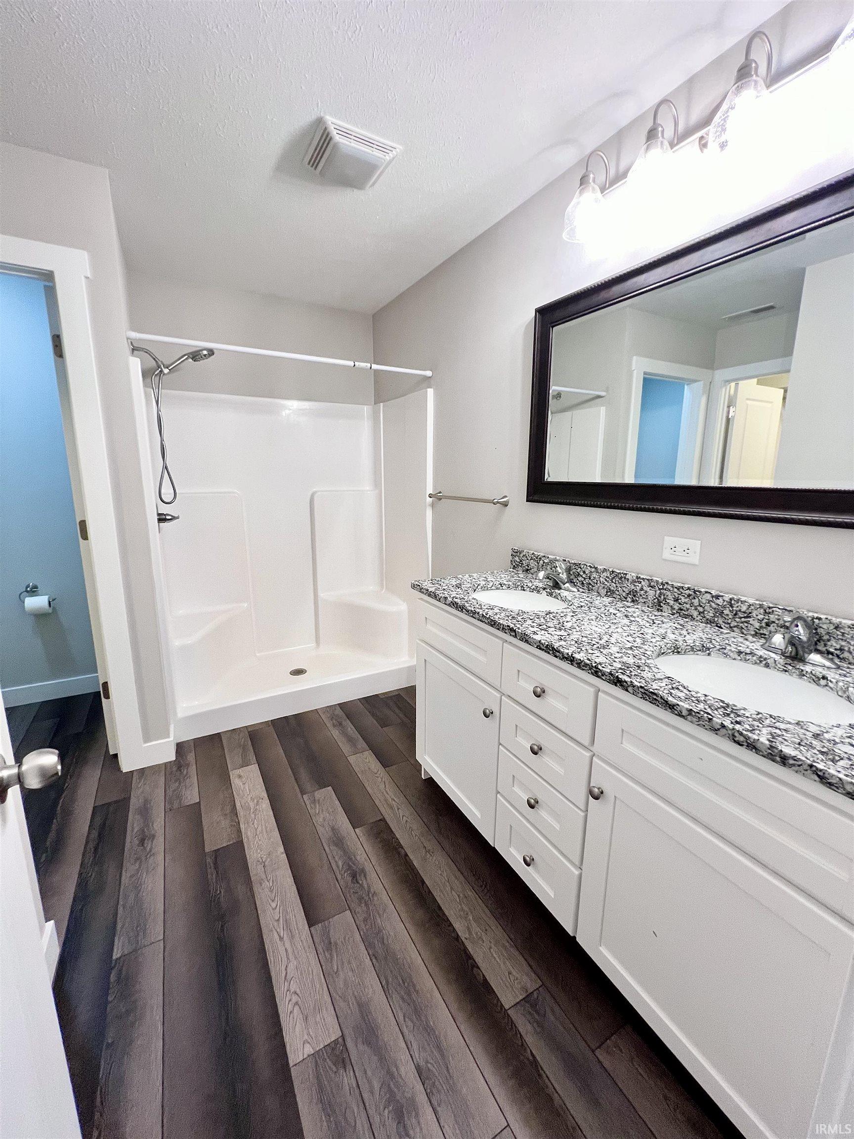 Wood-finish flooring, white double vanity with granite countertop, dual undermount sinks, and a walk-in shower