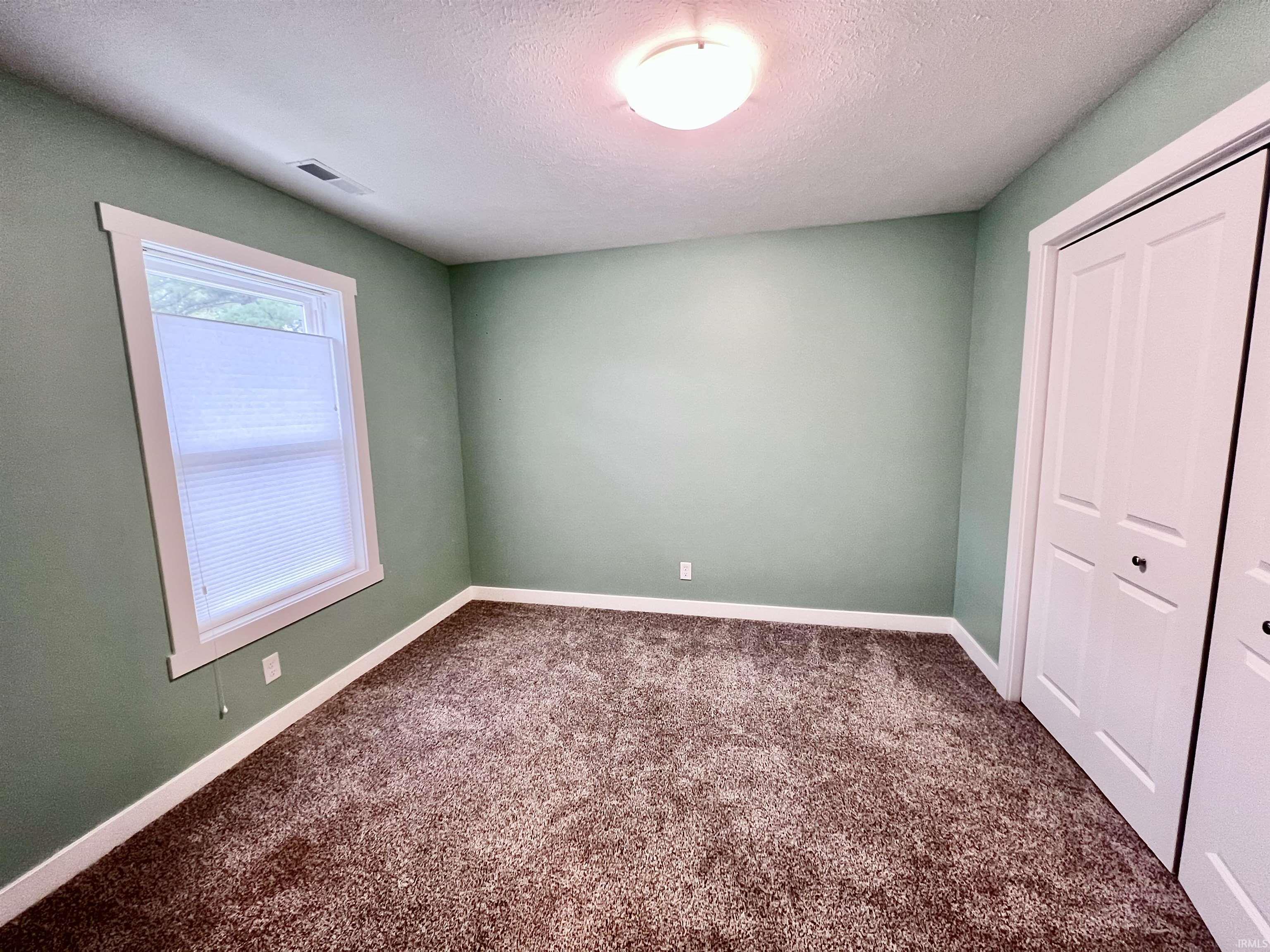 Spacious bedroom featuring soft sage-green walls, recessed ceiling lighting, large double-hung window with natural light, and a pair of white closet doors with panel detailing