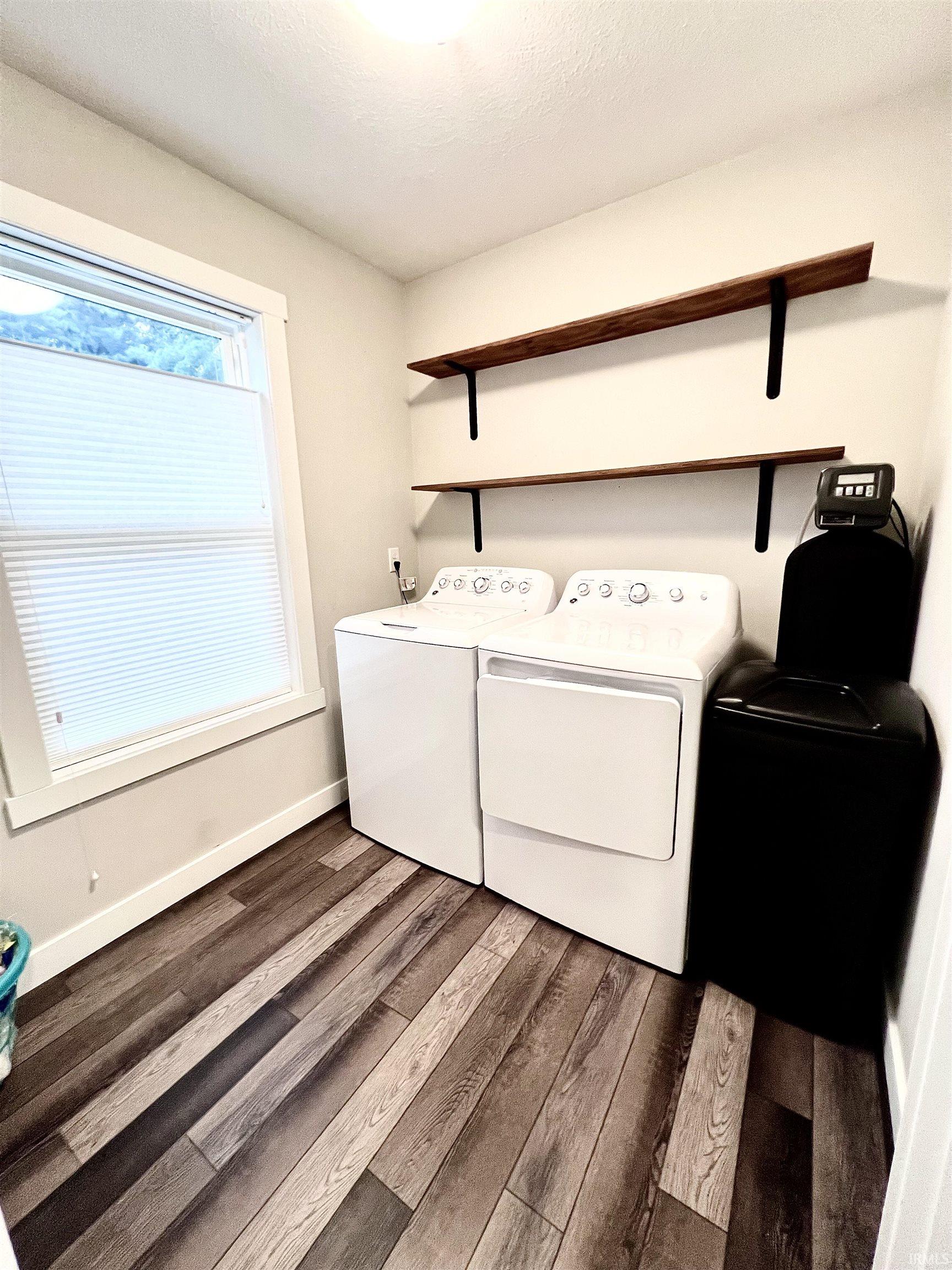 Dedicated laundry room featuring side-by-side washer and dryer appliances, wood-finish flooring, dual wooden floating shelves with metal brackets for storage, and a large window providing natural light