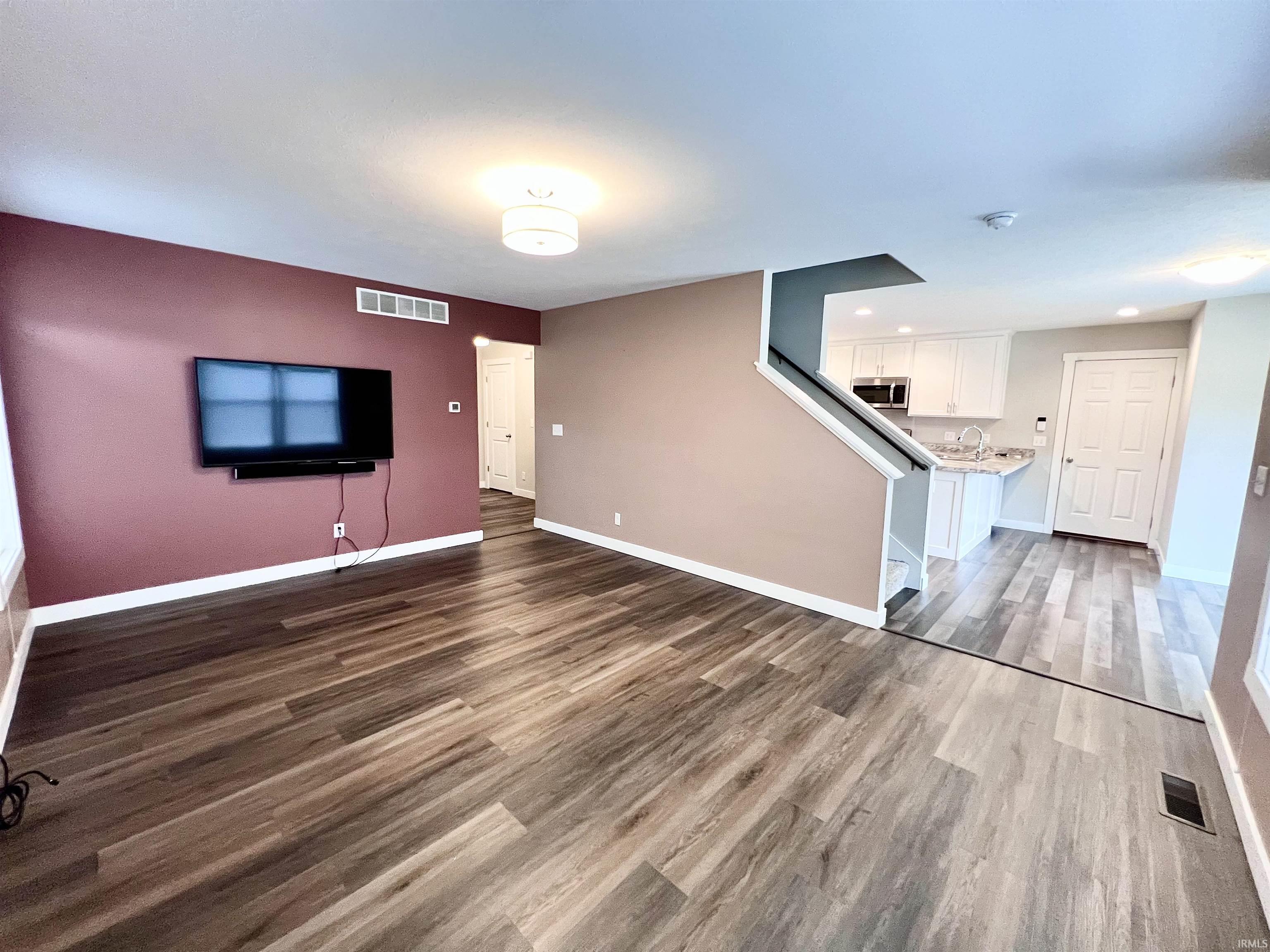 Spacious open-concept living area featuring rich wood-finish flooring, accent wall in deep burgundy, wall-mounted flat-screen television, recessed ceiling lighting, and architectural staircase with modern metal railing