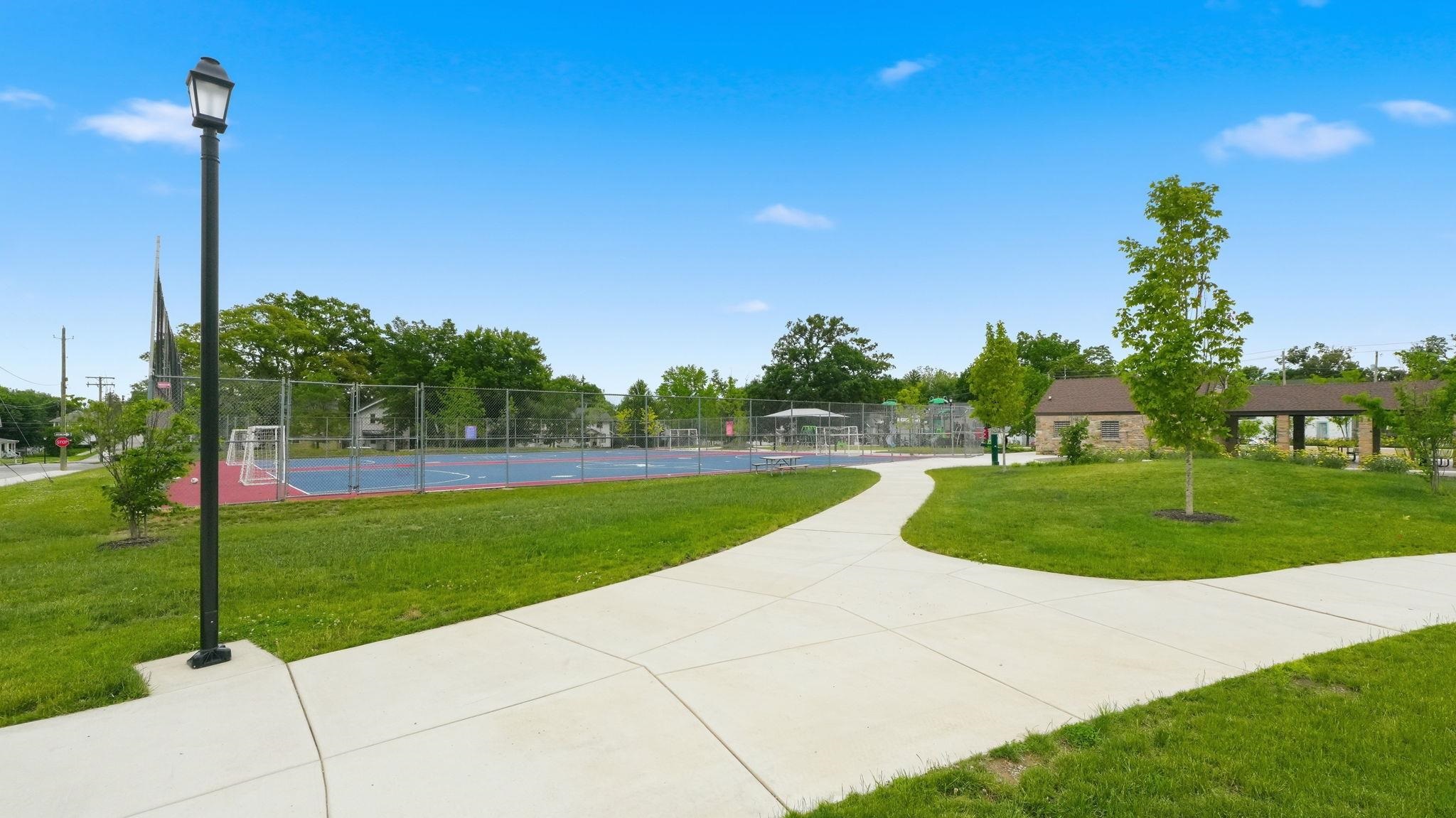 Paved walking paths featuring a street lamp, green lawn, fenced sport courts with goal posts, and a covered pavilion