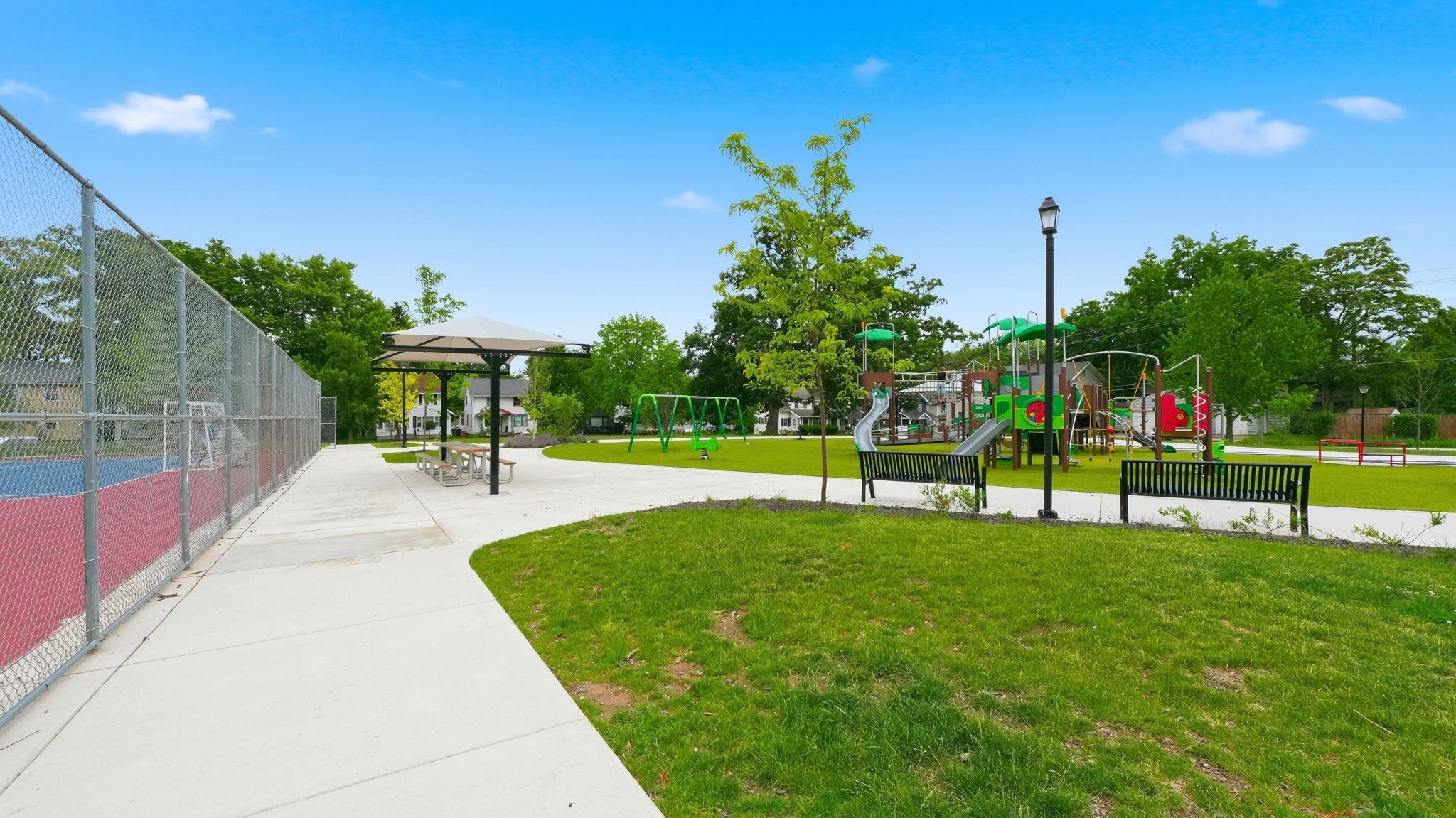 Community park featuring a fenced multi-sport court with red and blue surfacing, a play structure with slides and swings, paved walking paths, benches, and mature trees