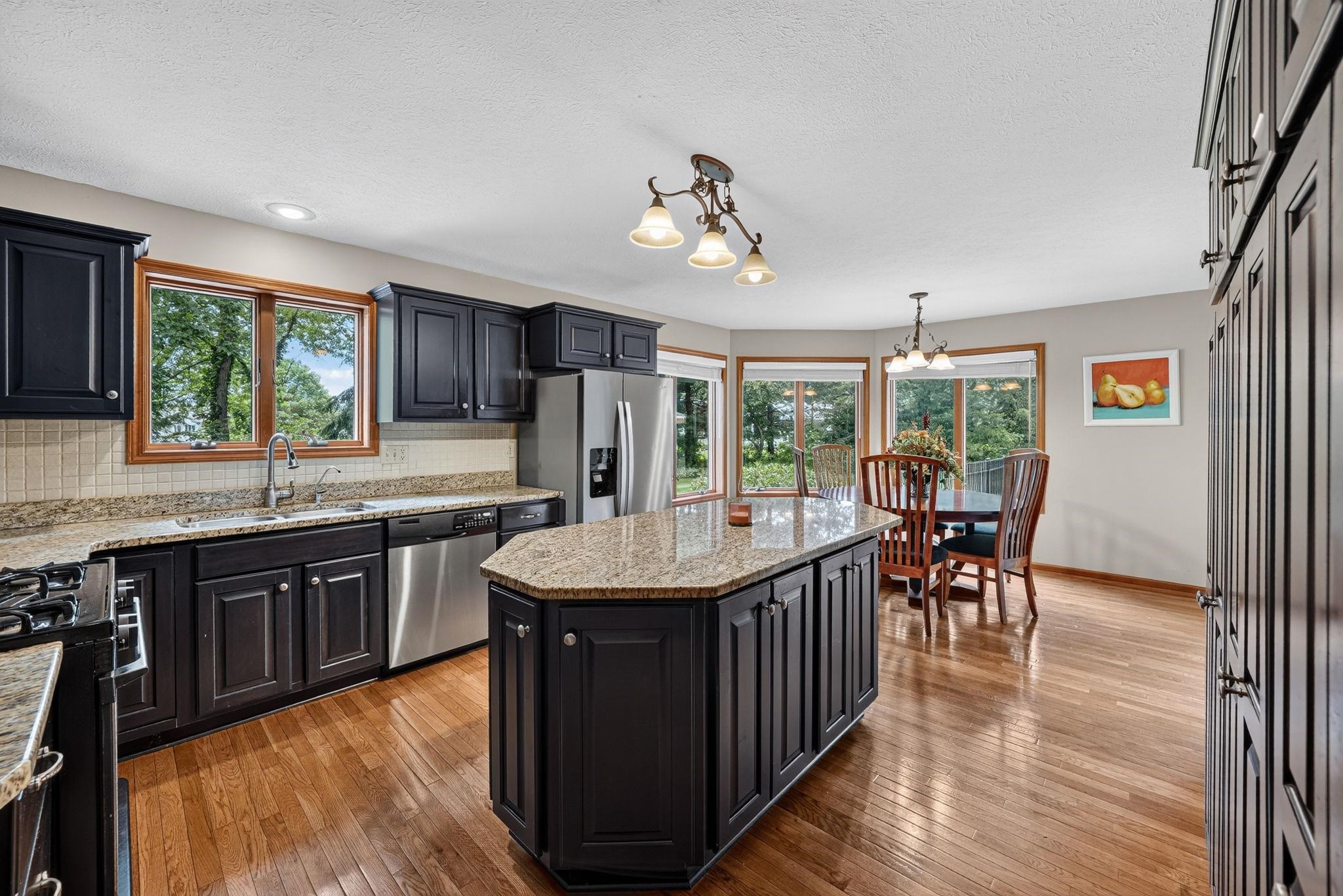 Kitchen featuring black cabinetry, stainless steel appliances, granite countertops, wood-finish flooring, and a dining area with a bay window