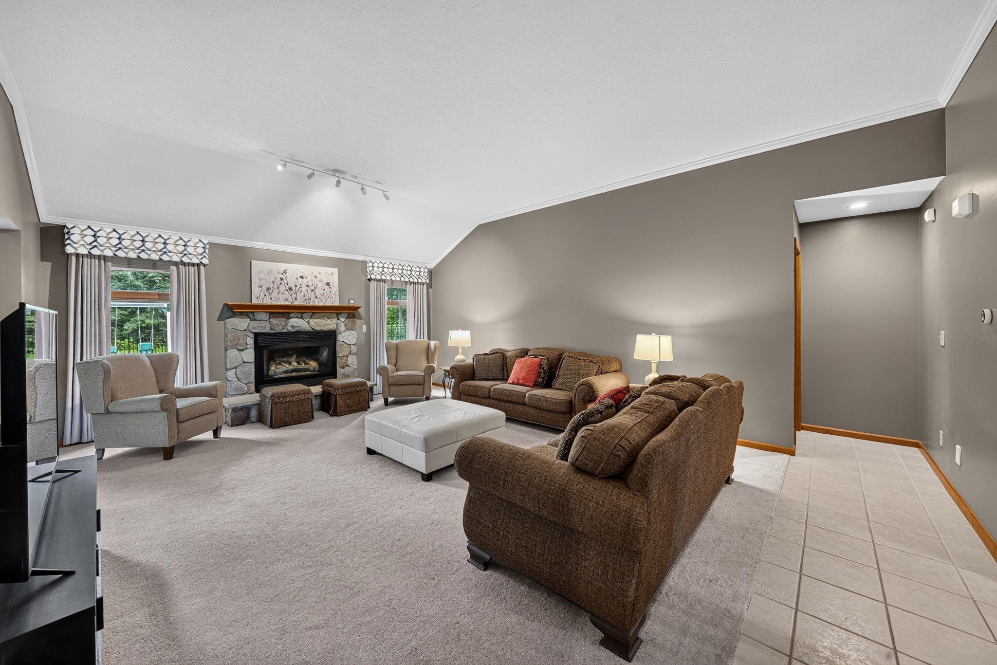Spacious living room featuring a stone-surround fireplace with a wood mantle, vaulted ceiling with track lighting, and soft neutral wall tones