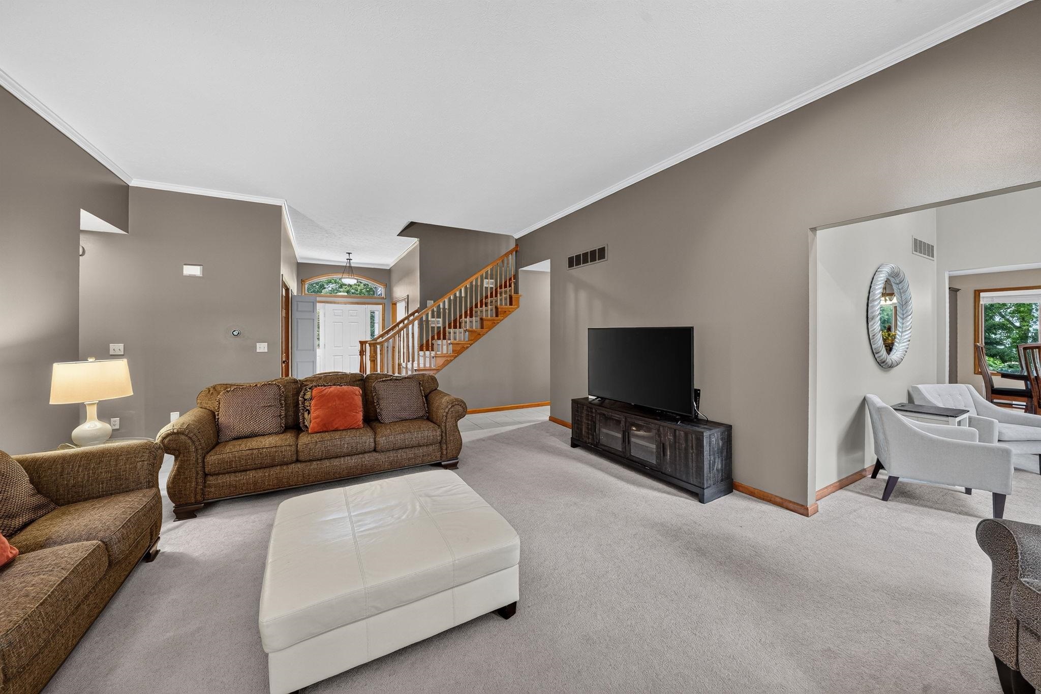 Spacious living room featuring high vaulted ceilings, light neutral carpeting, and a visible wood staircase with white risers