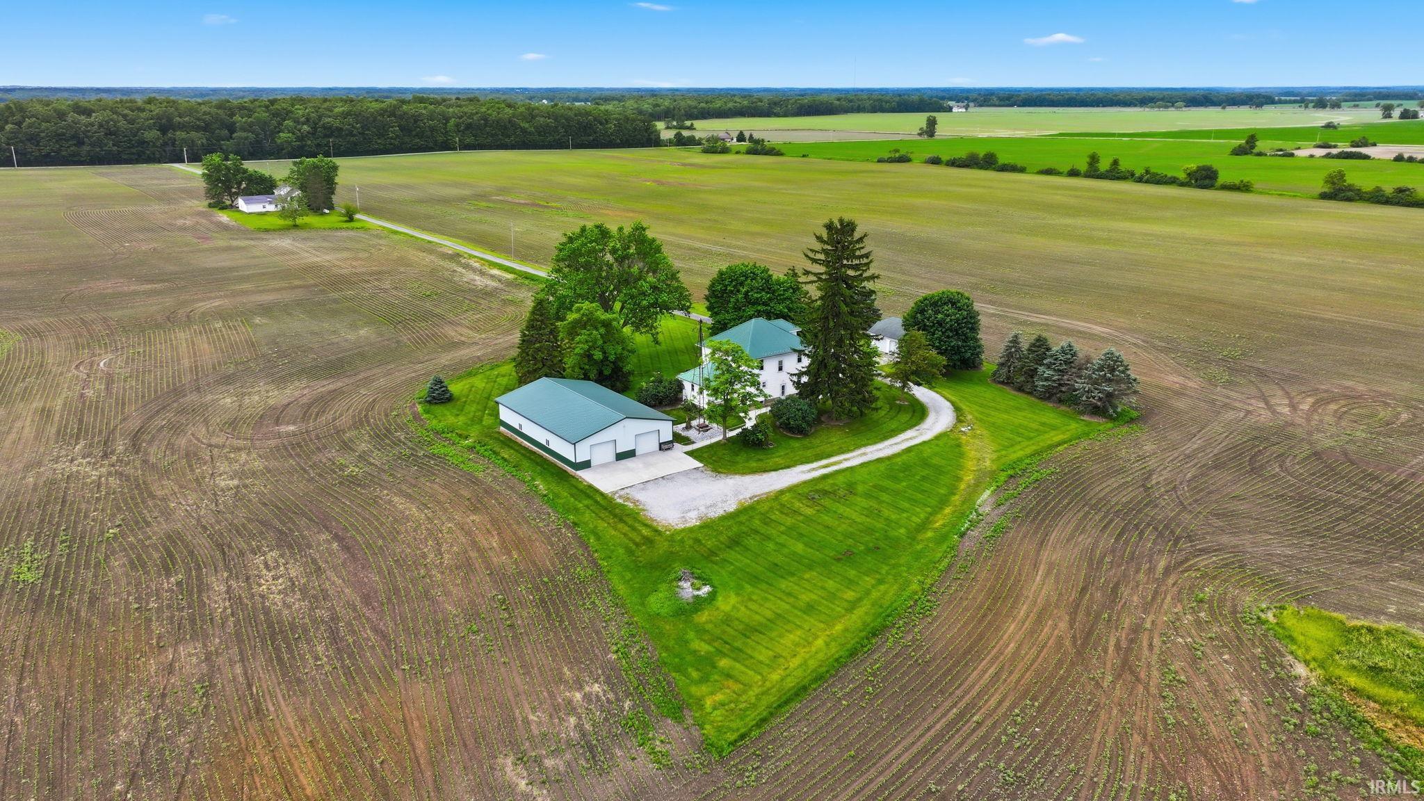 Aerial perspective showcasing a white residence with a green roof, accompanied by a large green-roofed outbuilding, situated amidst expansive cultivated fields