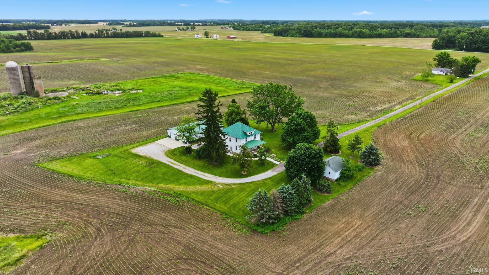 Expansive agricultural property featuring a white residence with a green roof, a circular driveway, mature trees, and outbuildings